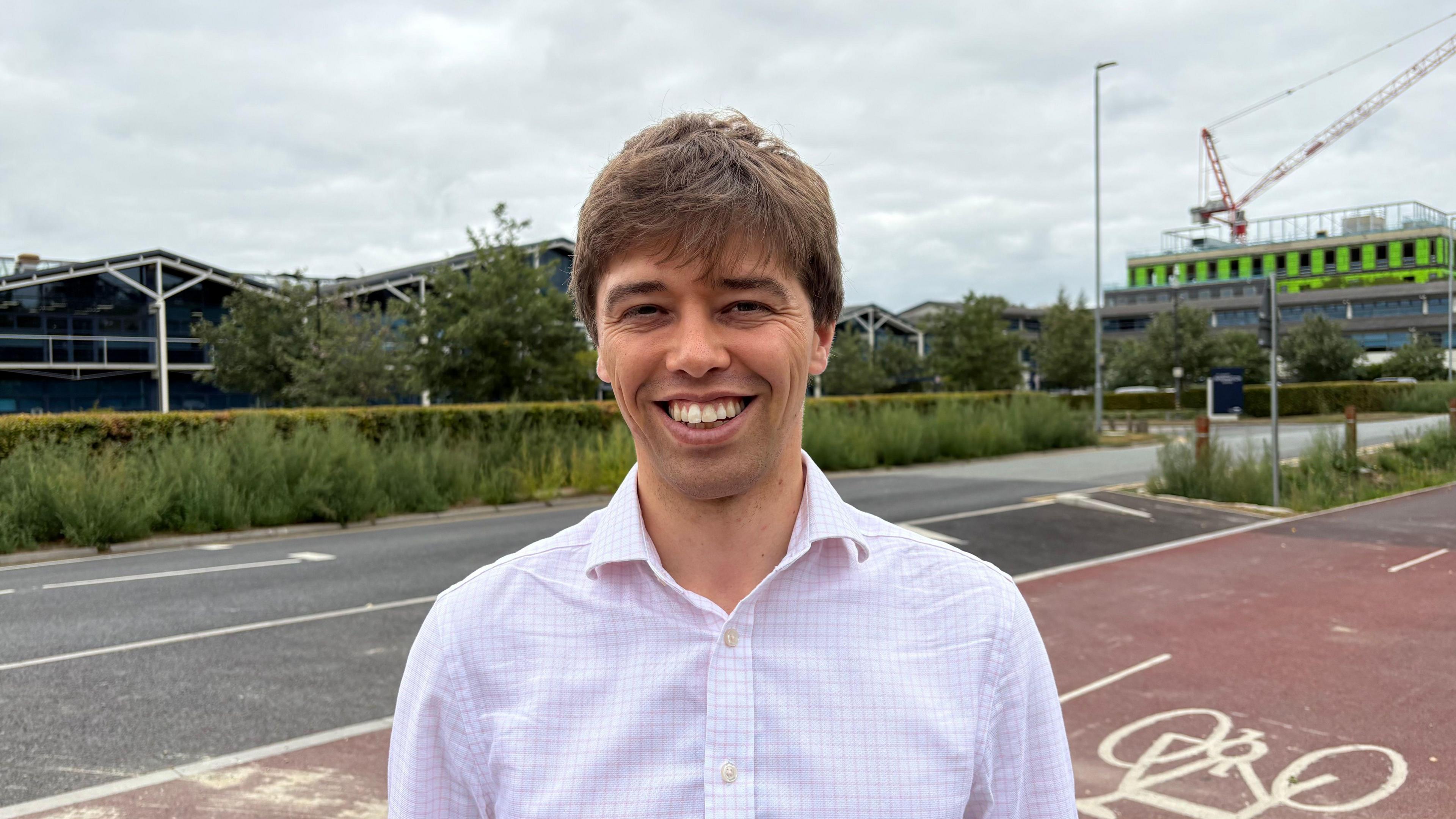 Cameron Holloway looking at the camera and smiling. He is wearing a light coloured shirt. Behind him is a road and cycle path, as well as buildings and a crane.