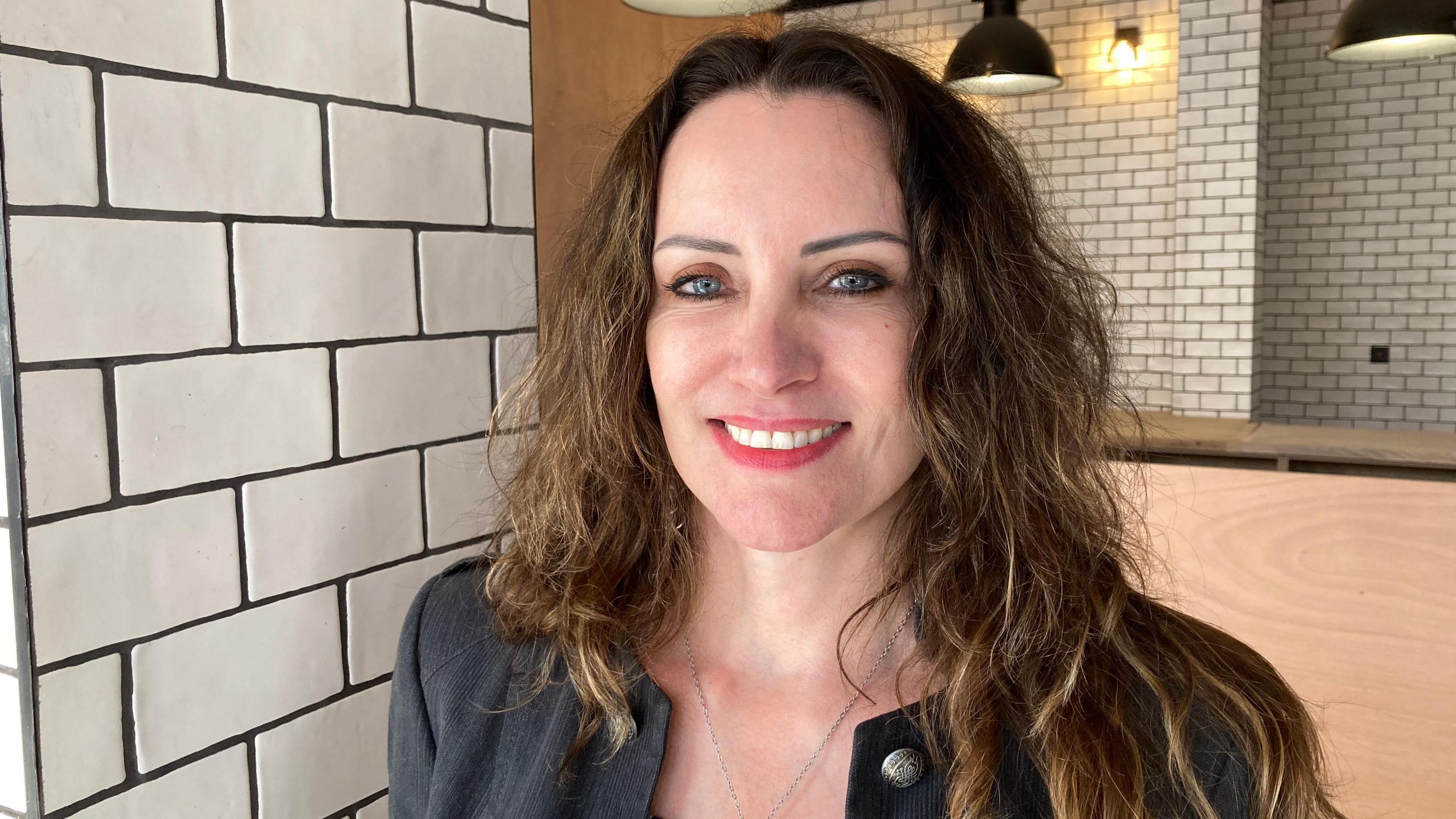 Suzanne Thompson standing in the Key Cafe, smiling at the camera. She has long, wavy brown hair and piercing blue eyes. She is wearing a dark grey jacket and a silver necklace chain. Behind her are various columns and walls covered in white tiles with black grout. 