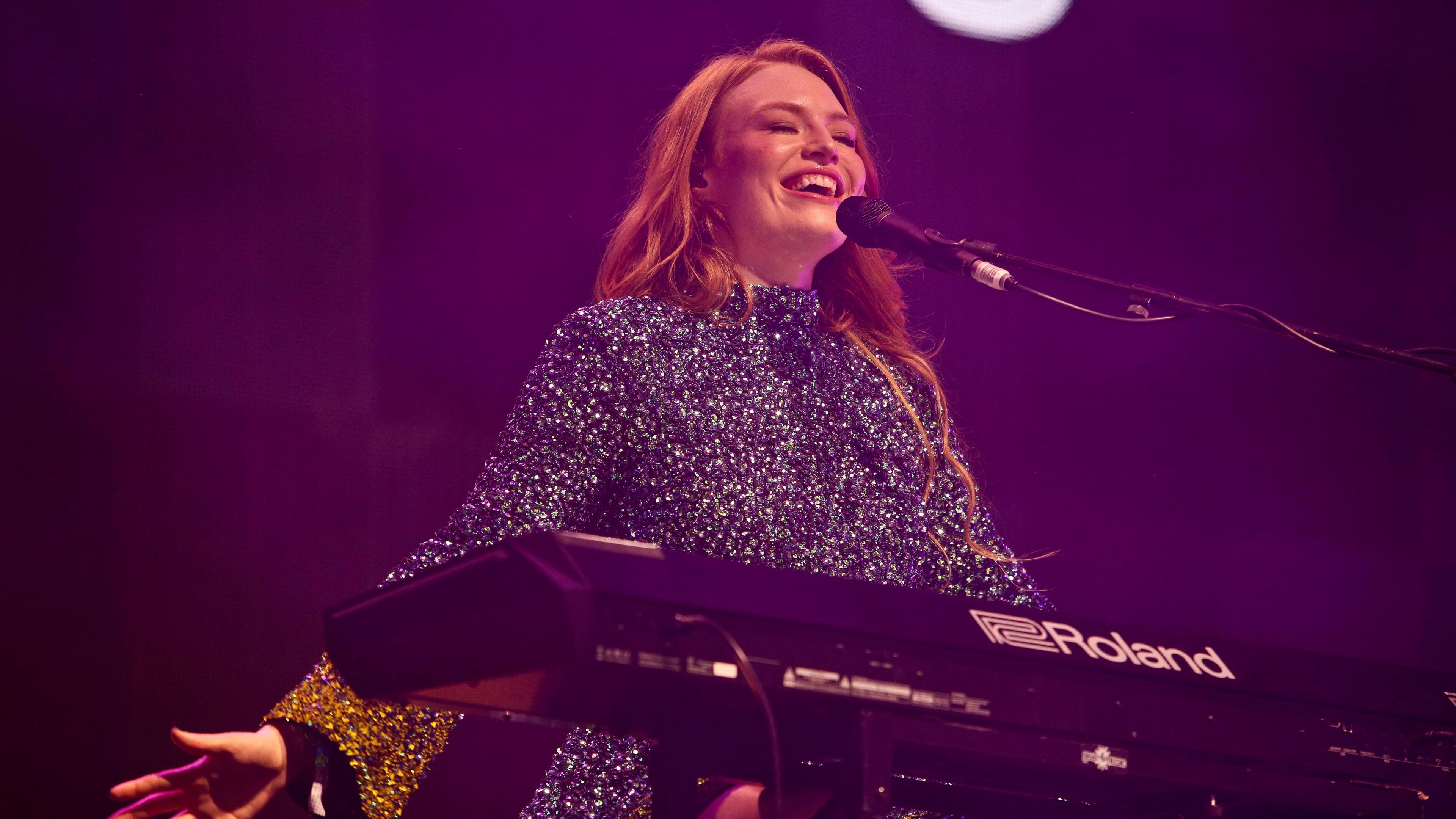 Freya Ridings in a sparkly dress sat behind a keyboard and microphone. She has her arms out to her side and is smiling while singing with her eyes closed. She has long ginger hair.