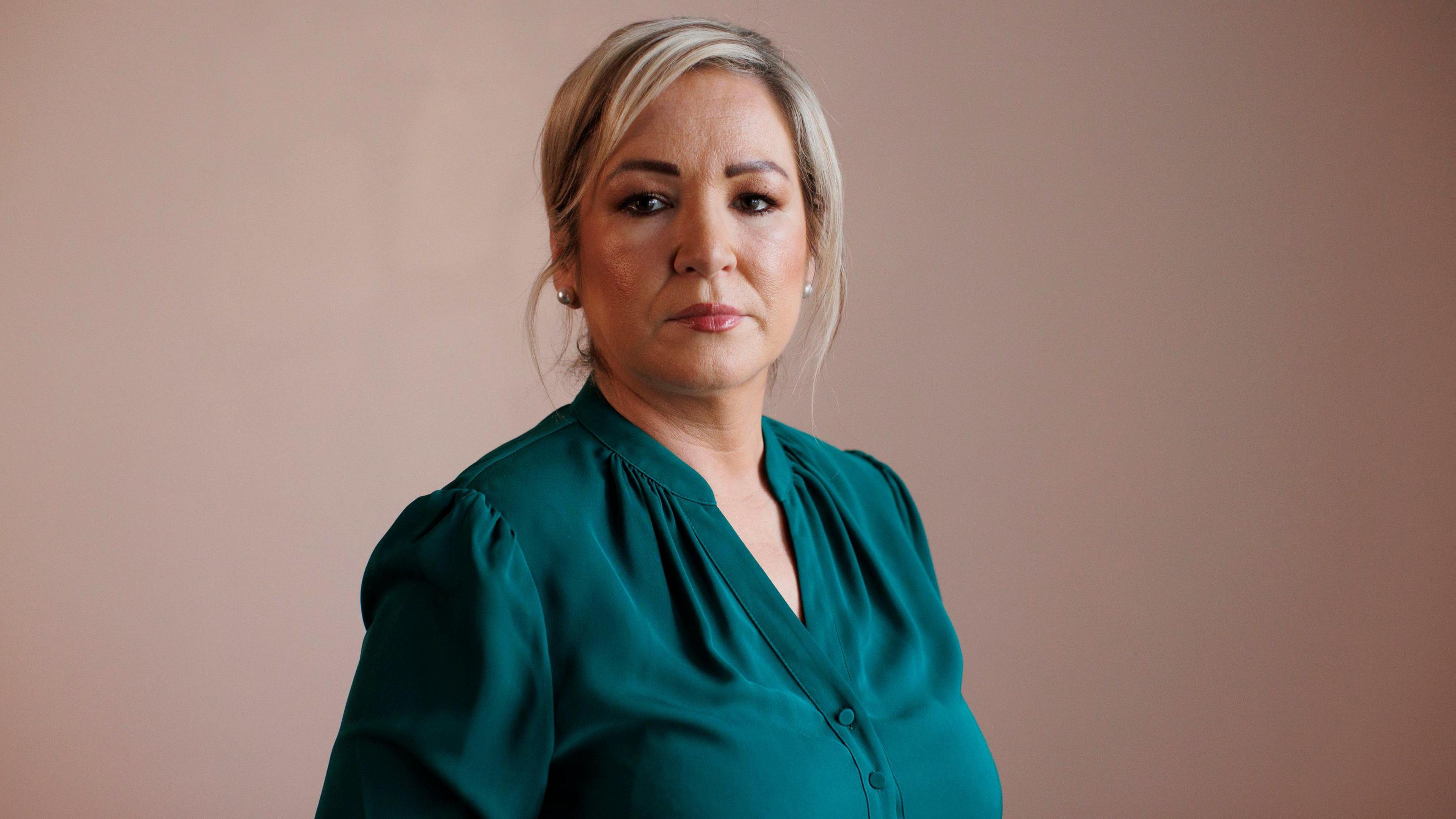 Michelle O'Neill looking to camera with a serious expression.   She has blonde hair tied back and is wearing earrings, lipstick and a dark green blouse.