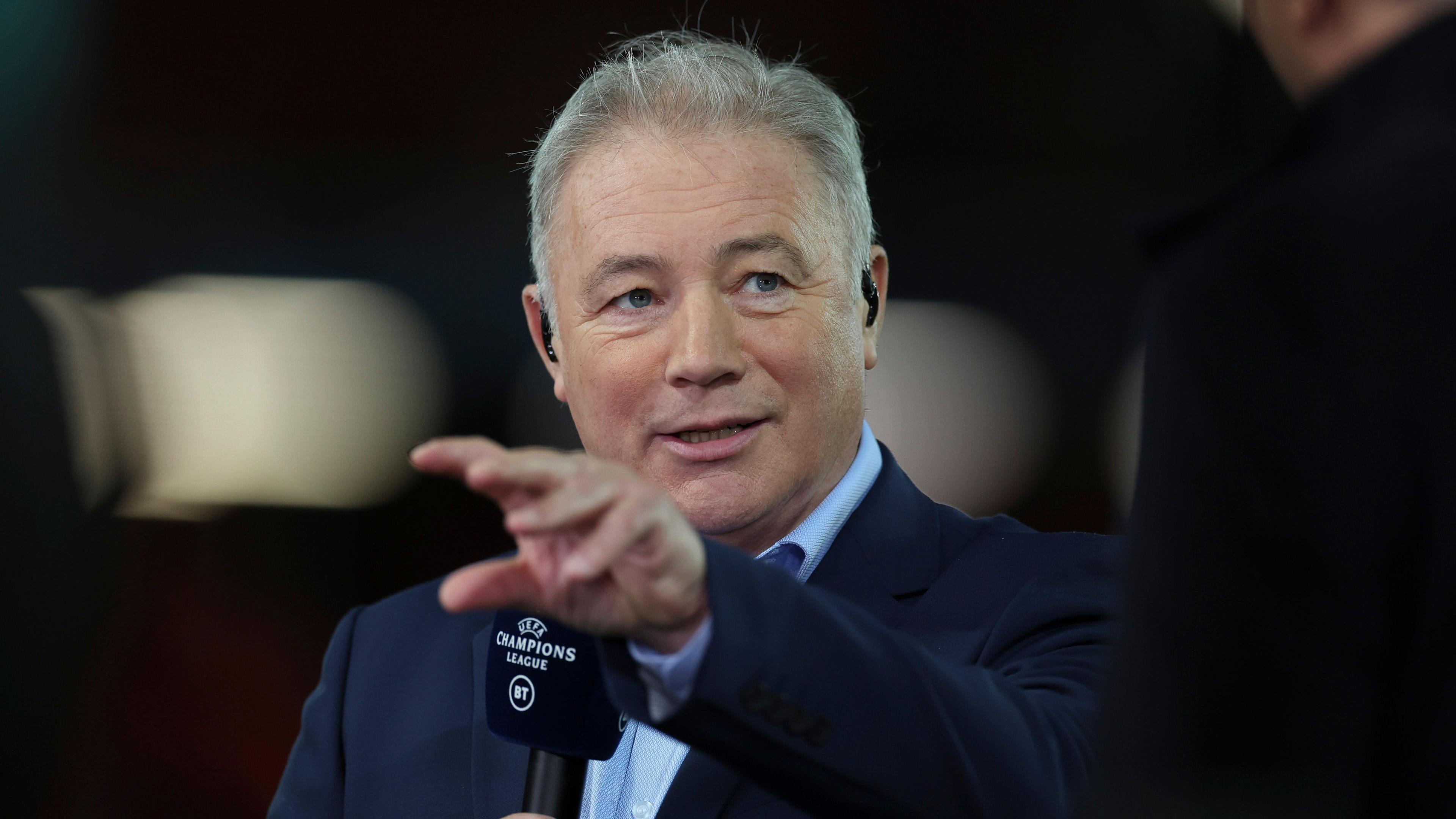 Ally McCoist is holding a microphone and talking into it at a sport event. He is moving his hands in front of his face while he is talking.