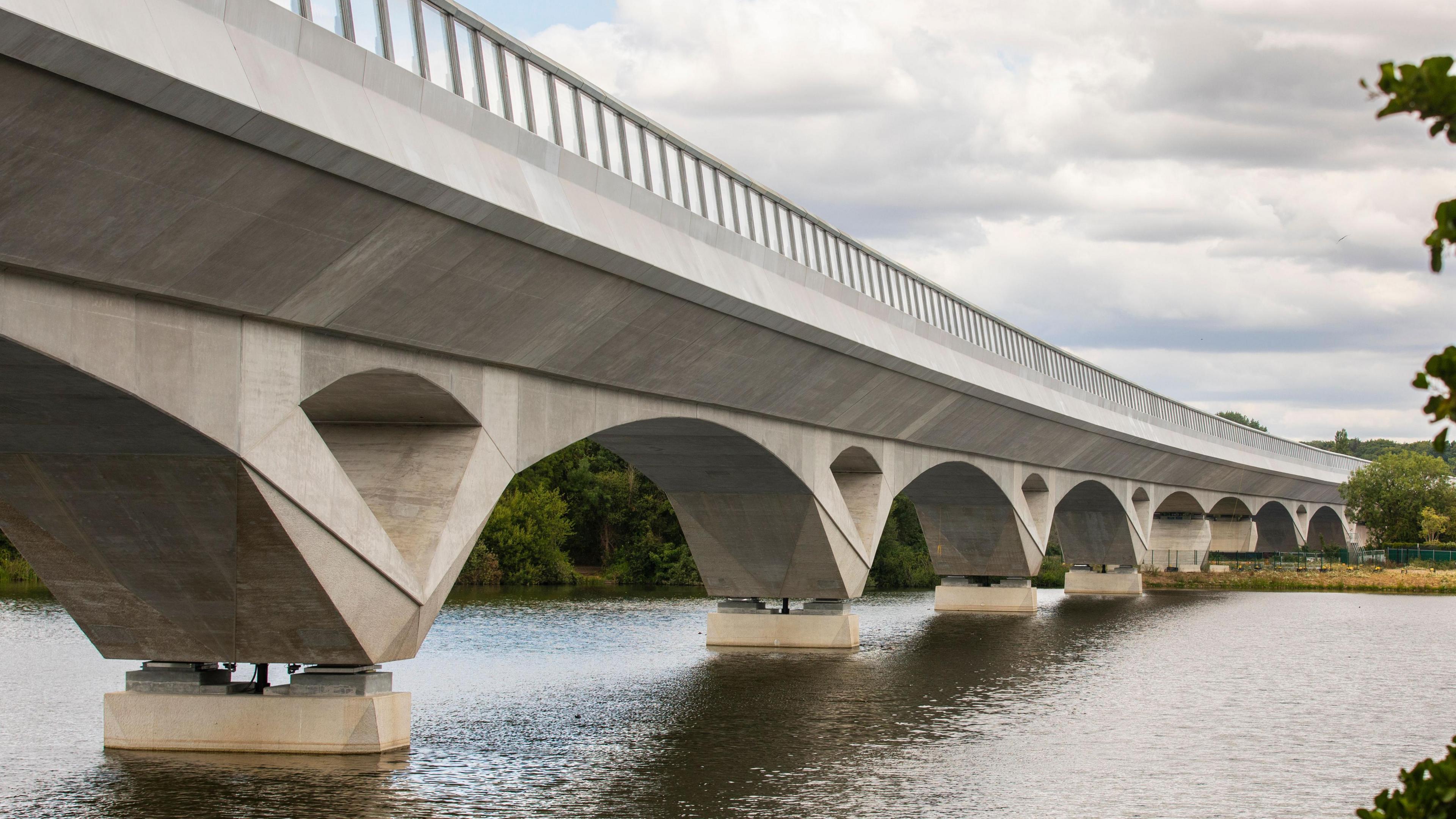 A white bridge is standing on top of water. It runs from the left of the image to the right which is on land. There are trees in the background.