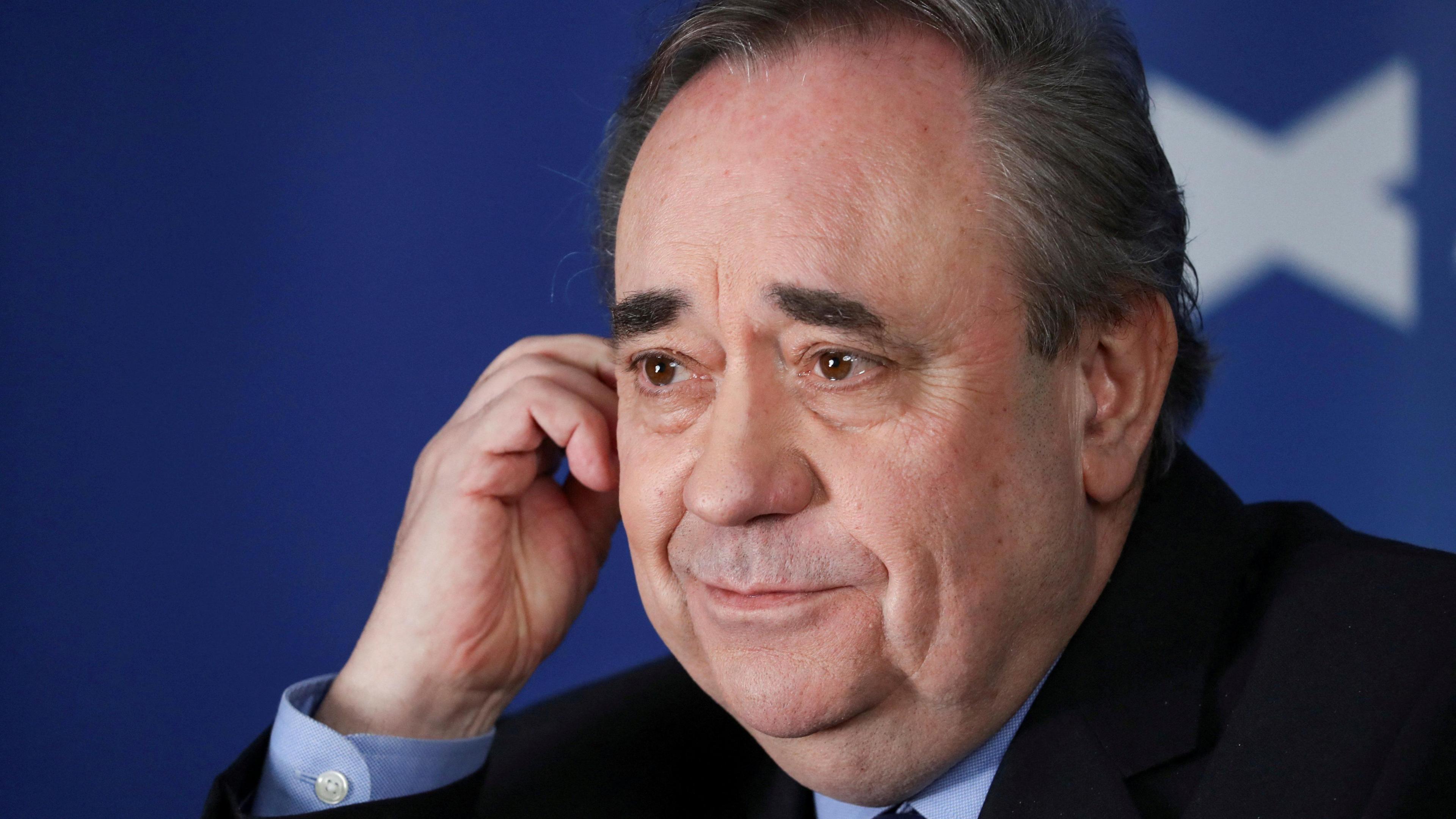 Alex Salmond in close up against a saltire background. He is wearing a dark jacket, mid-blue shirt, and is smiling.