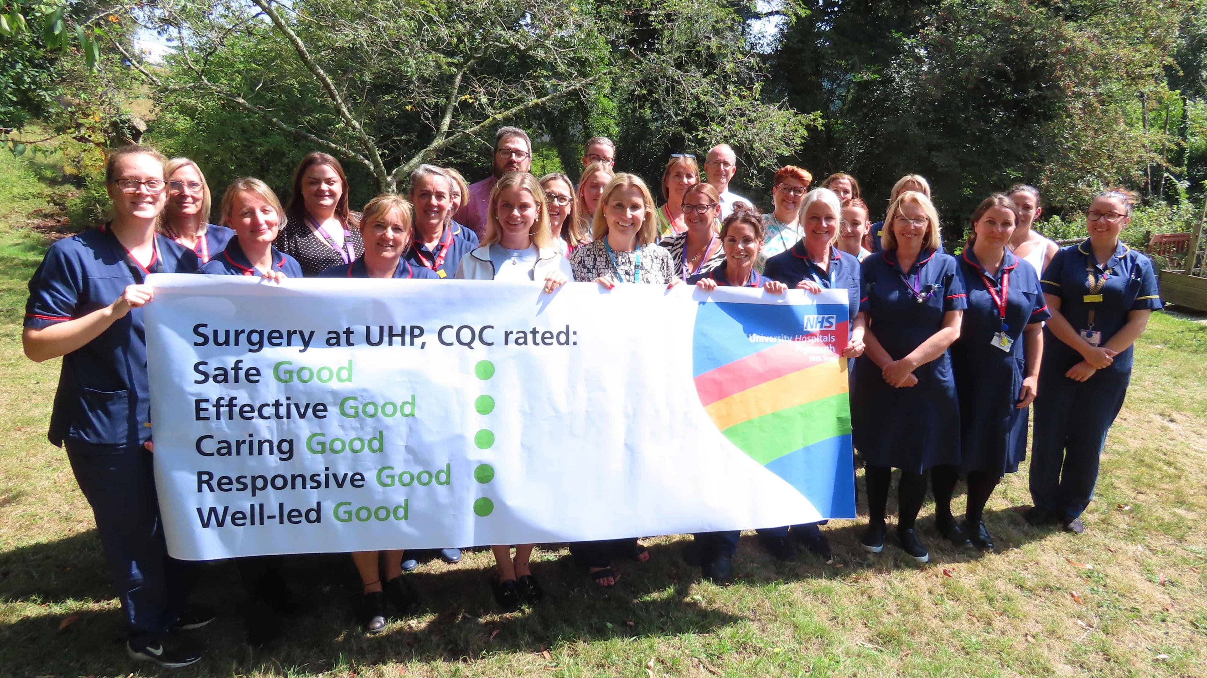 Staff from Derriford Hospital stand on a lawn in front of trees and hold up a banner saying CQC has rated surgery services at the site as good. The word good is written five times in green along with five green dots. Many of the staff are wearing scrubs.