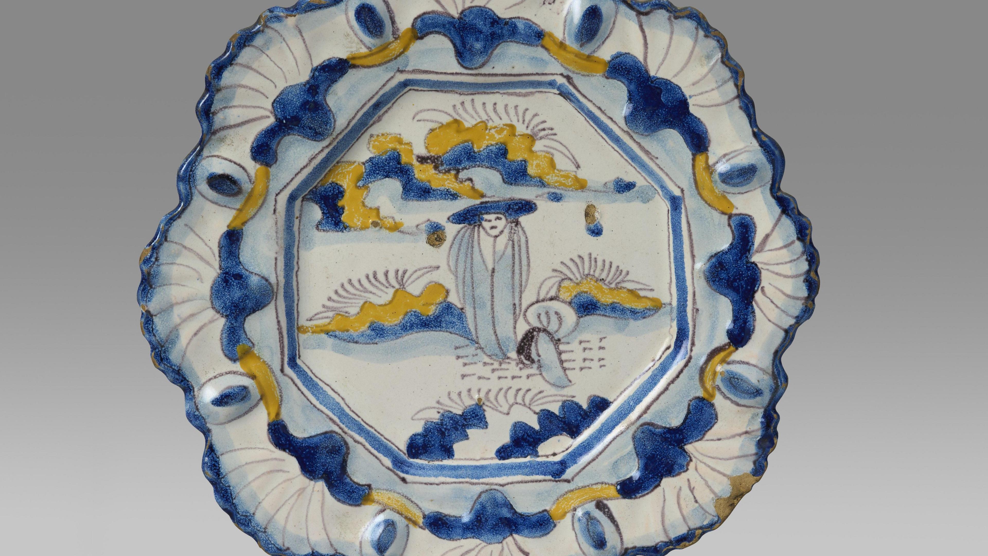 The rare yellow, blue and white plate shows a Chinese figure standing in a rocky landscape. The plate is against a white backdrop.  The rim of the plate is moulding with a repeating shell motif.