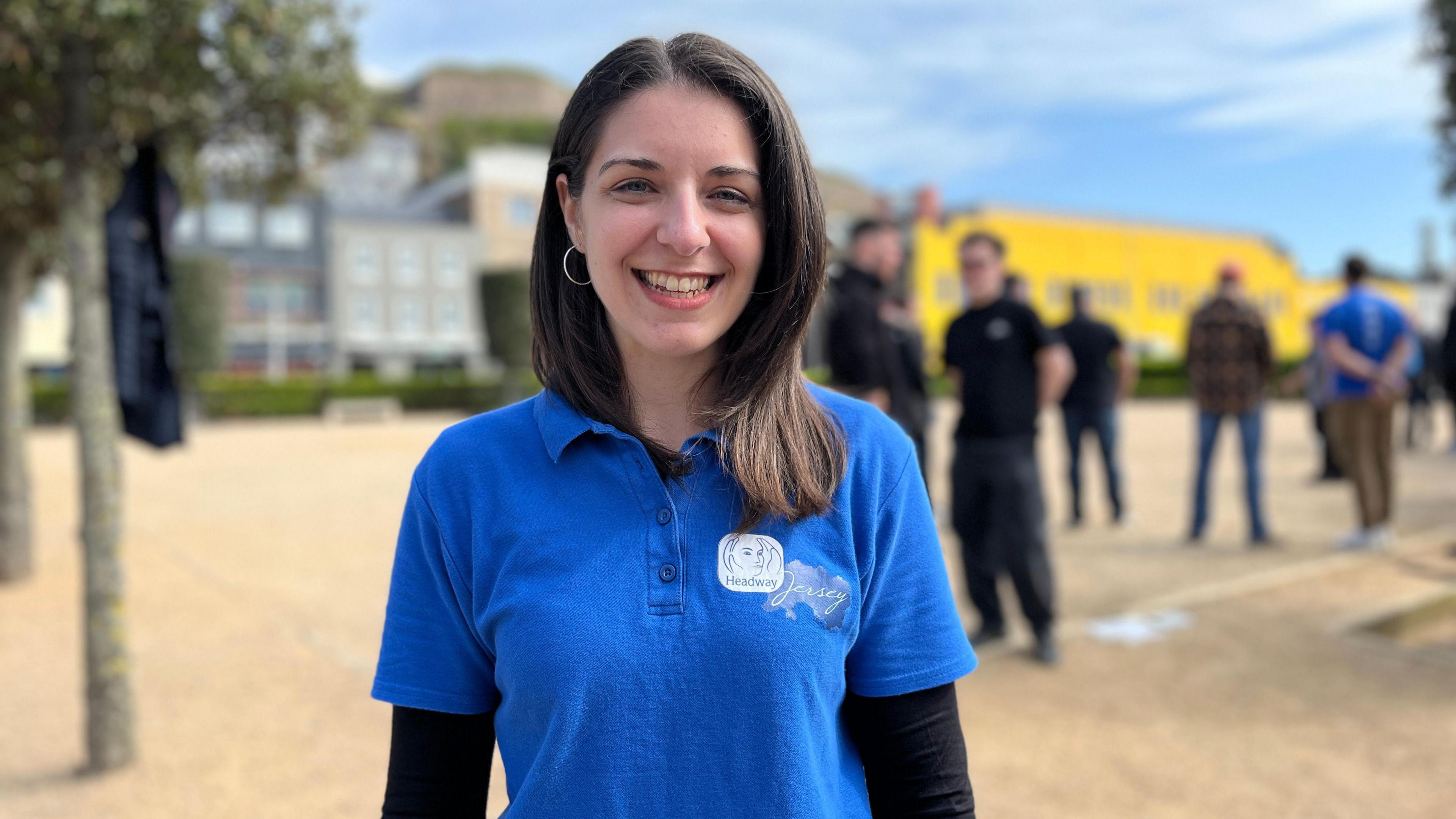 Sarah smiles at the camera wearing a blue Headway Jersey top and in the background teams stand by waiting to play pétanque