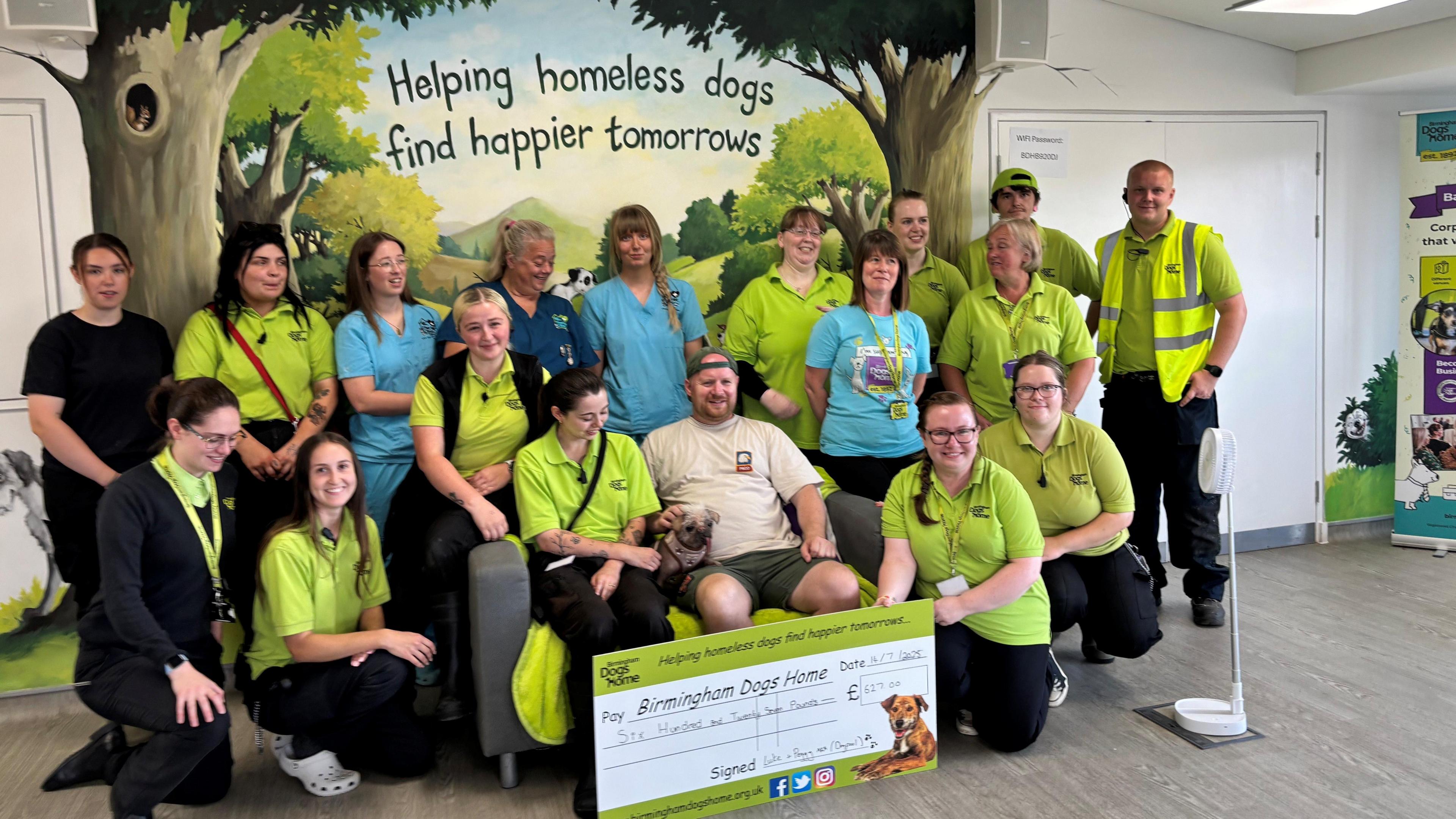 Peggy – a pug and Chinese crested cross dog - sits on a sofa with her owner and is surrounded by a group of 17 volunteers from Birmingham Dogs Home, most of whom wear light green charity shirts. They are sitting and standing in front of a wall decoration reading "Helping homeless dogs find happier tomorrows"