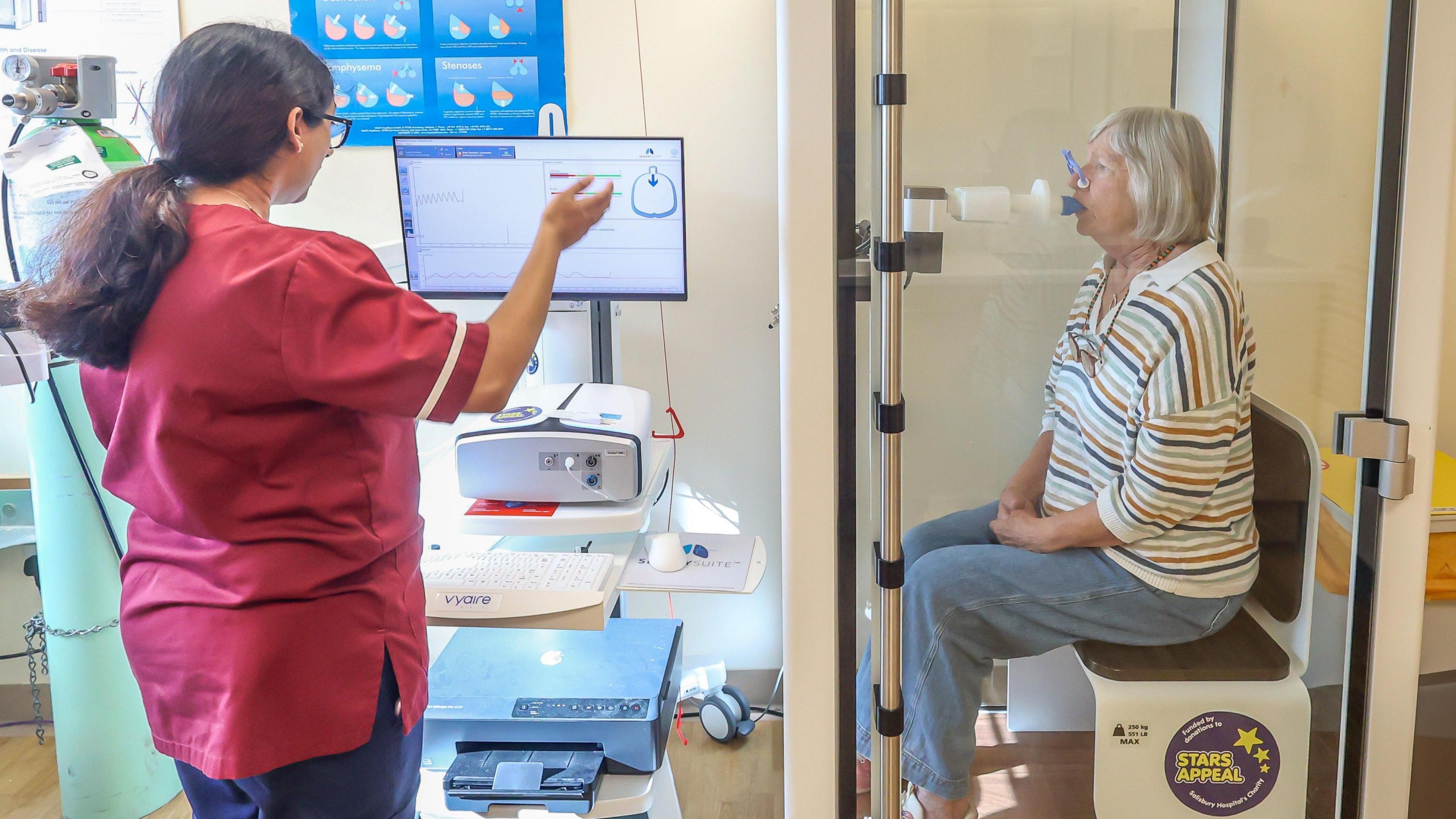 Patient Kirsty Dudin sits in a booth and breathes into a lung function testing machine. She is sitting on a plastic seat which says "Stars Appeal". Outside the booth, a doctor monitors the feedback from a computer.