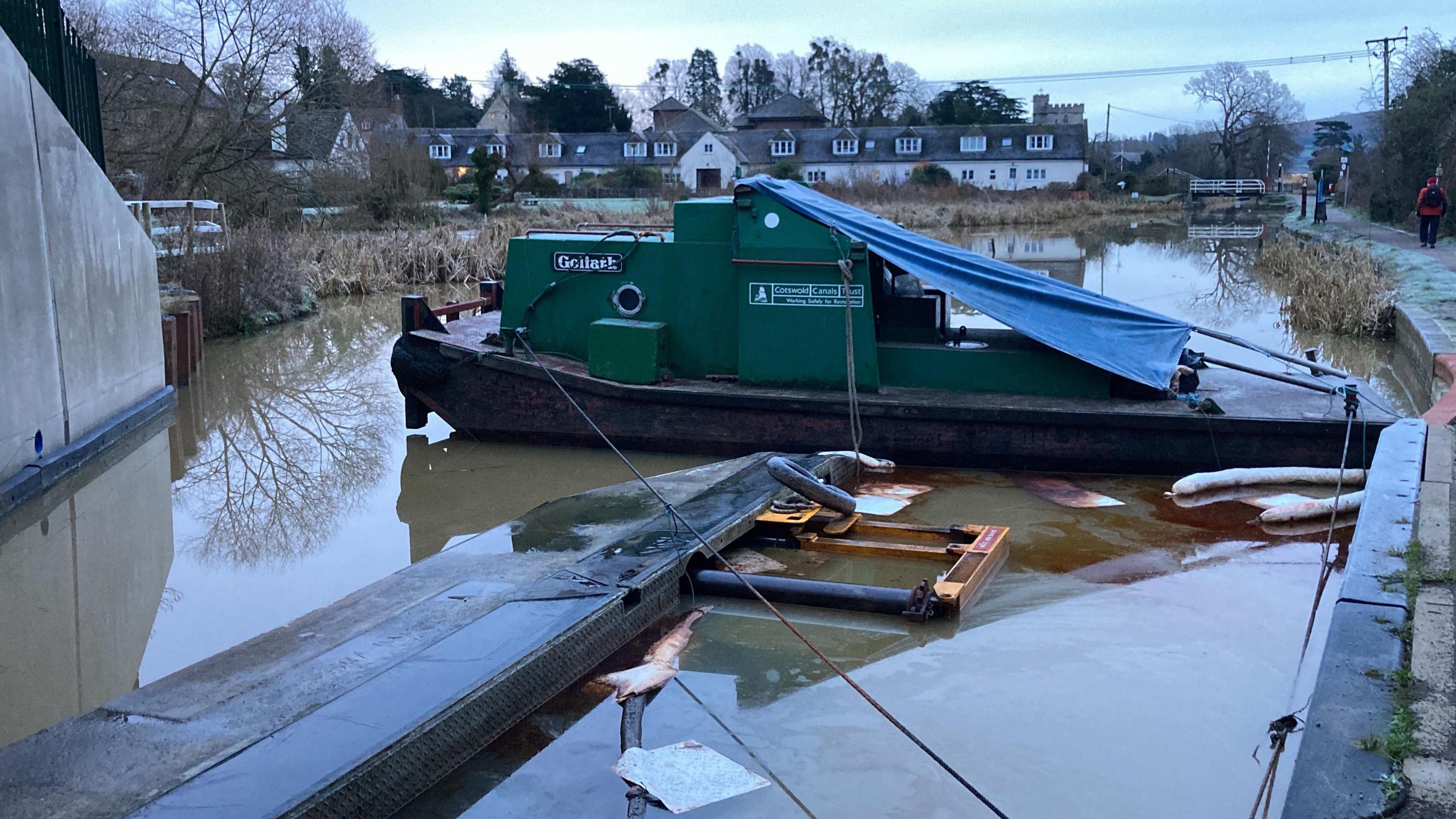 A sunken dredger barge on a canal on a cold winter's day. There are houses in the background, and people can be seen walking along the frosty towpath next to the canal. There is a large oil spill beside the barge.