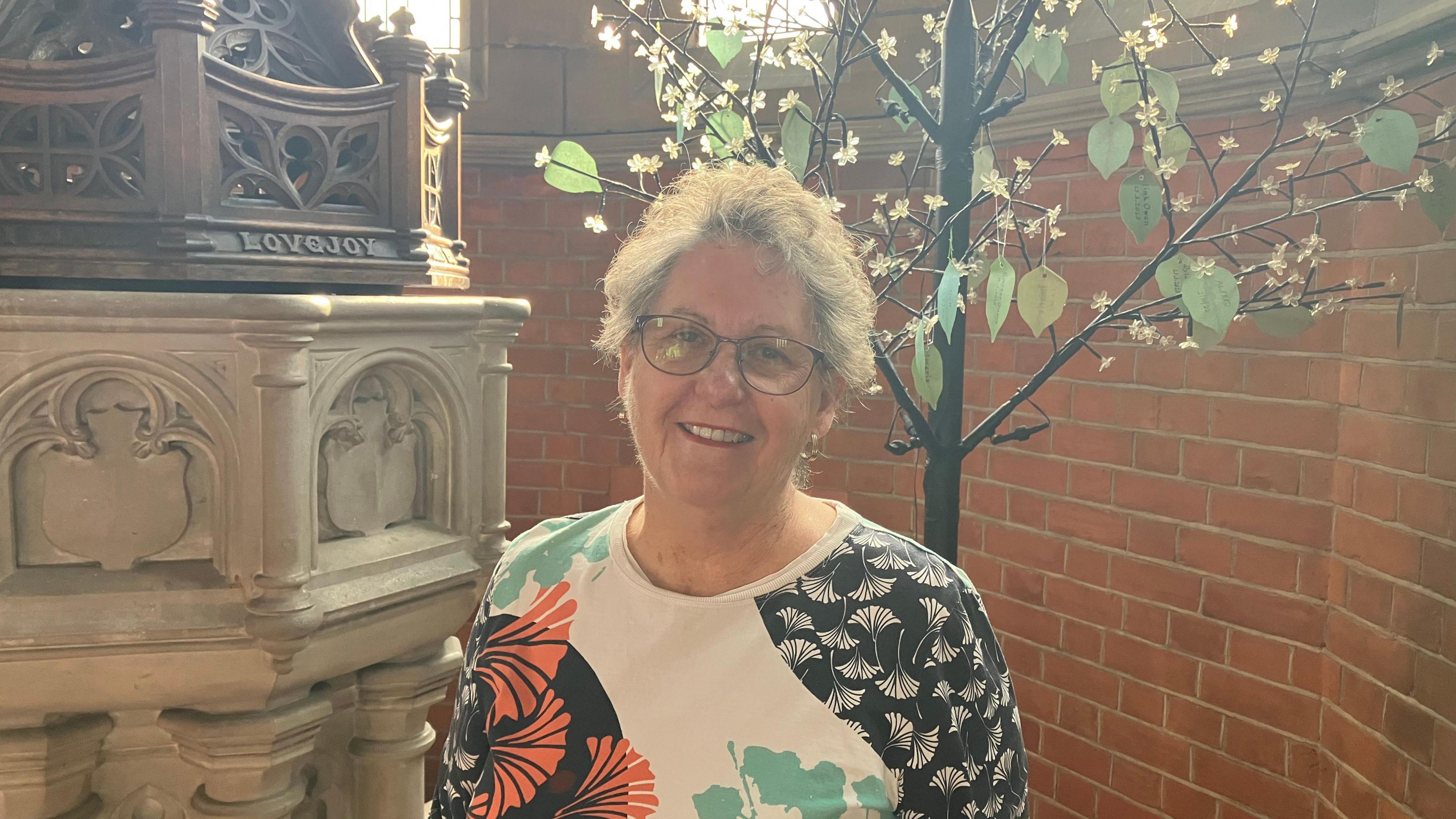 Wendy Grantham looks at the camera and smiles. She is wearing a white top with a floral pattern in white, orange and black and has short pale-coloured hair. Behind her is a church font and the brick wall of the church with a fake tree adorned with green leaves.