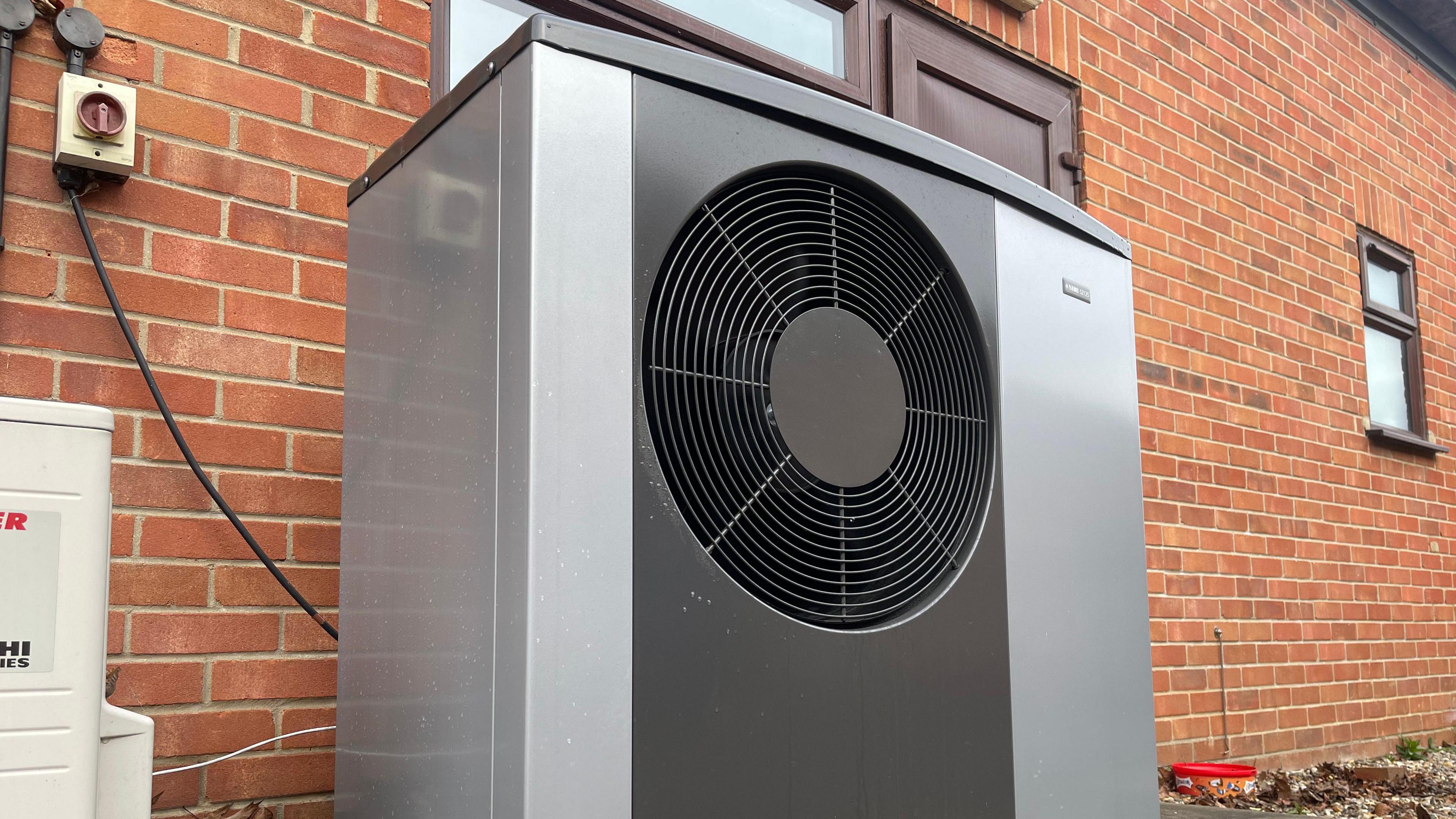 A grey heat pump against a redbrick wall on a gravel surface.  