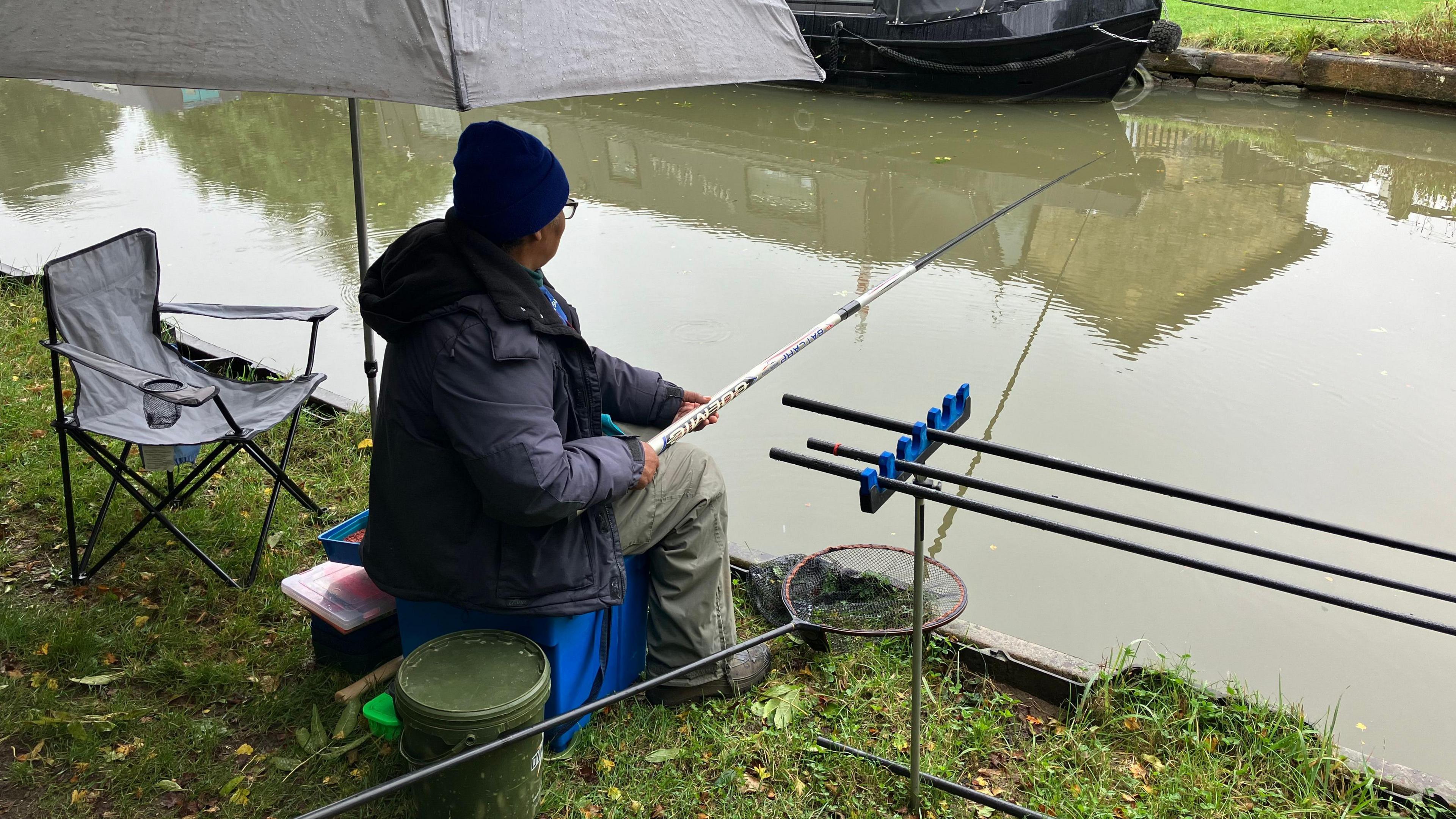 A woman in a navy coat and blue woolly hat sits under an umbrella on a blue box holding a 3 metre fishing rod into the canal