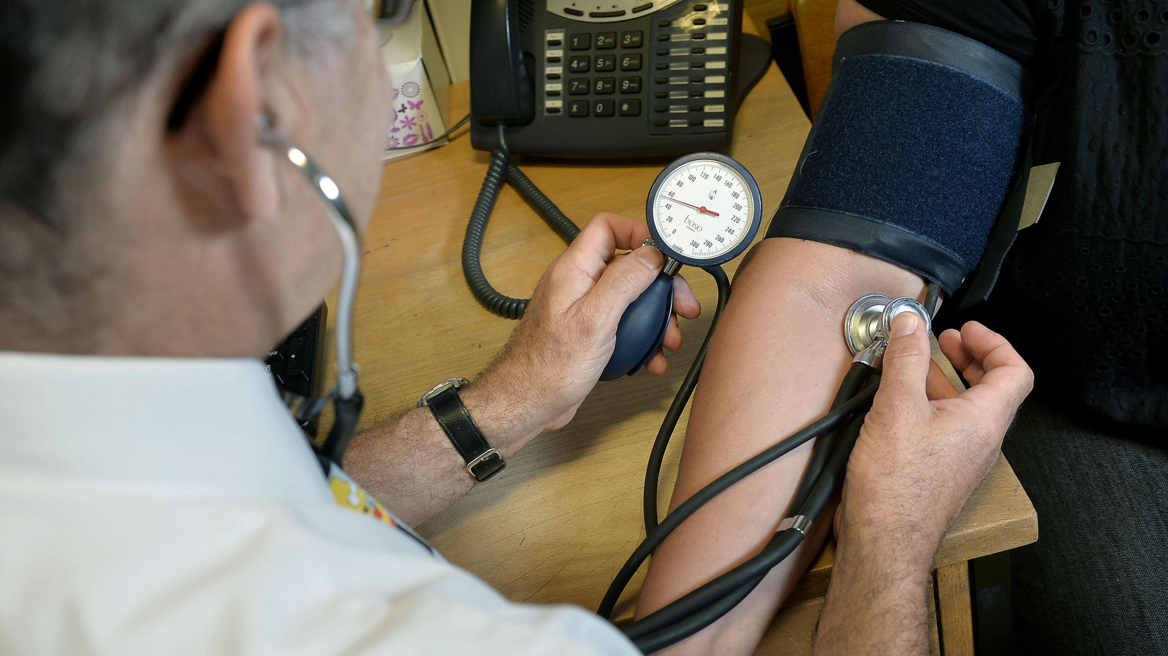 A GP is checking a patients blood pressure. The patient has a dark blue strap around their arm and the doctor is holding a small metal device just below the strap to monitor their heart rate. The doctor is to the left of shot, the back of his head can be seen. He is wearing a white shirt and is listening to the patients' heart rate. The photo was taken from over his shoulder.