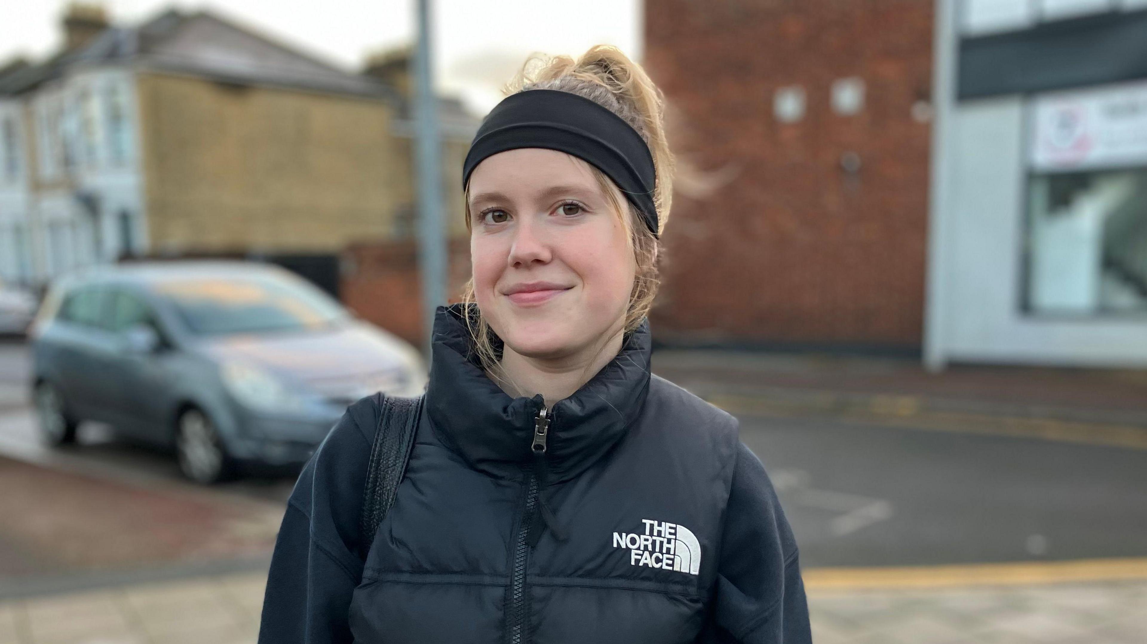 A young woman with blonde hair smiling at the camera. She is wearing a black headband and a black jacket and standing in front of some shops and a parked car.