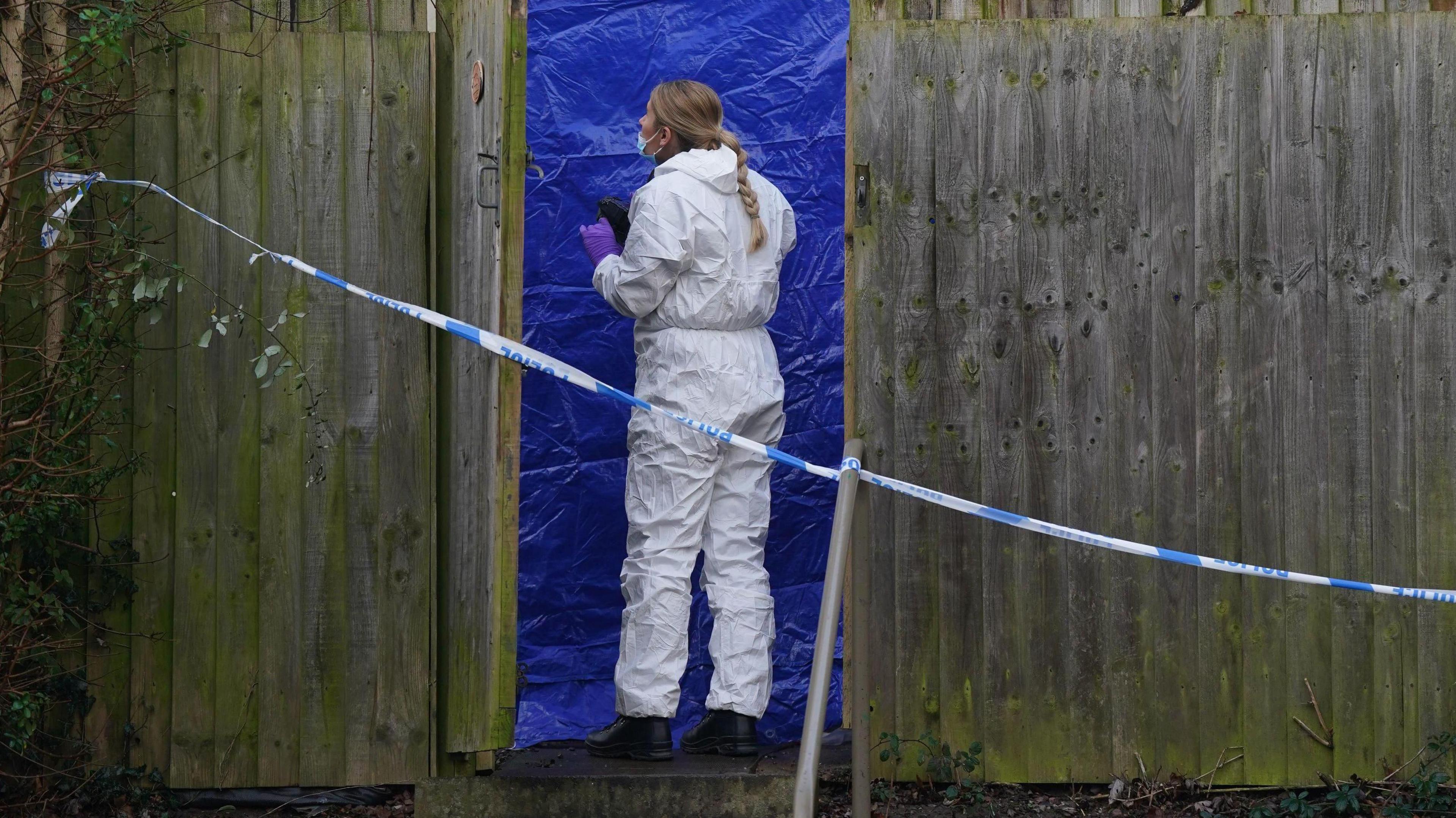 A forensic officer wearing white overalls standing in front of a blue tent with green fencing either side and blue and white police tape across the whole scene