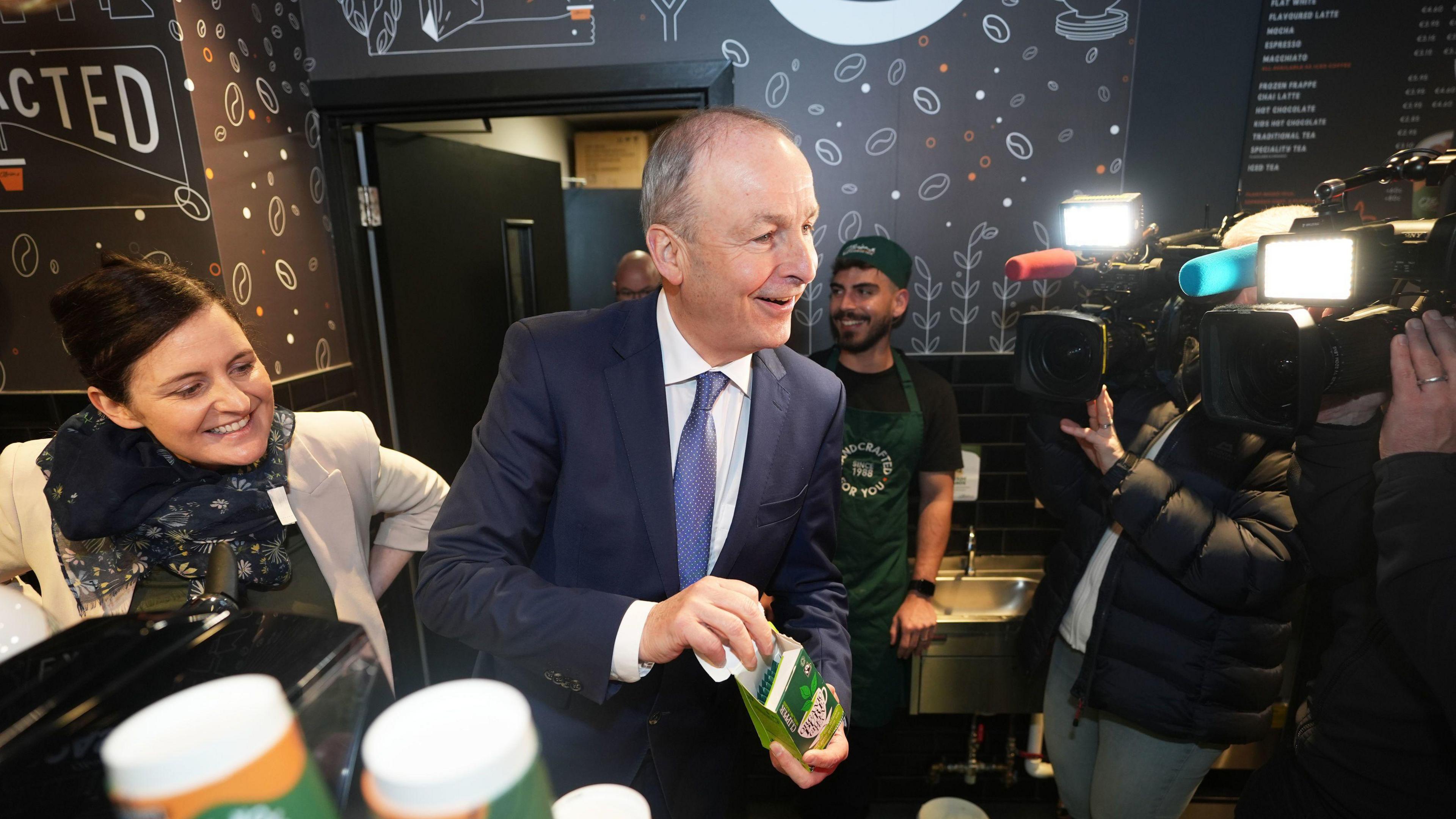 The photo is taken inside a coffee shop. In the middle of the photo is Tánaiste Micheál Martin, wearing a white shirt, blue tie and navy suit. He is looking at a camera crew, who are to the right of the photo. To the left of Micheál Martin is a woman, wearing a scarf and cream jacket. 