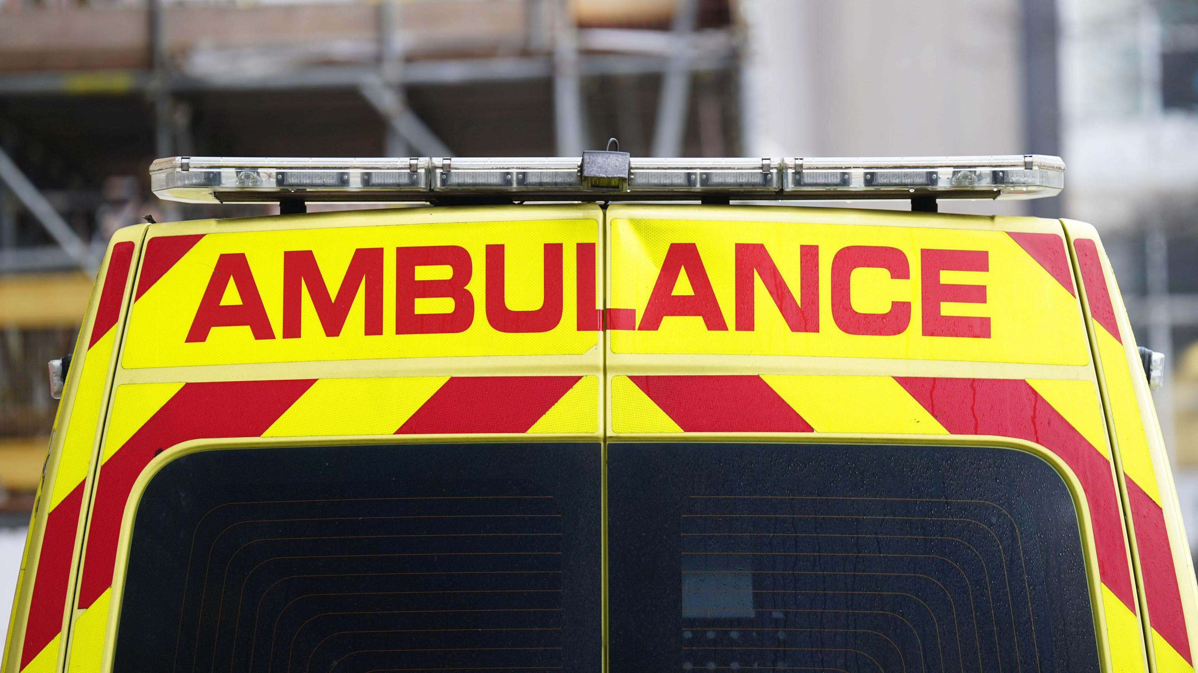 A picture of the back of an ambulance. It has yellow and red stripes. There is, in red writing, the word AMBULANCE. There is the lights attached to the top of the vehicle.