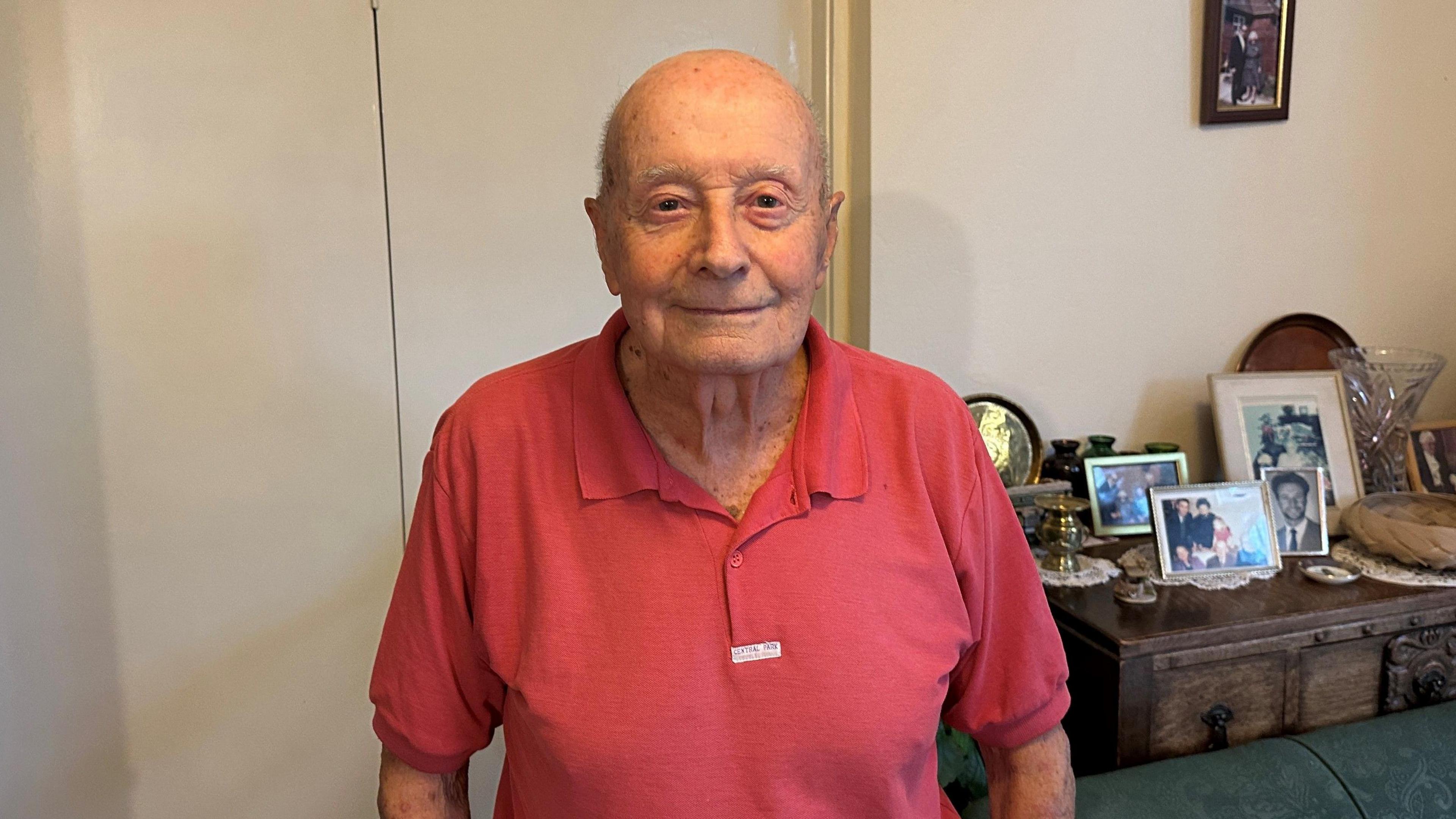 Peter Taylor, who is now 90, poses in a red t-shirt for a photo in his living room in front of family ornaments and photos.
