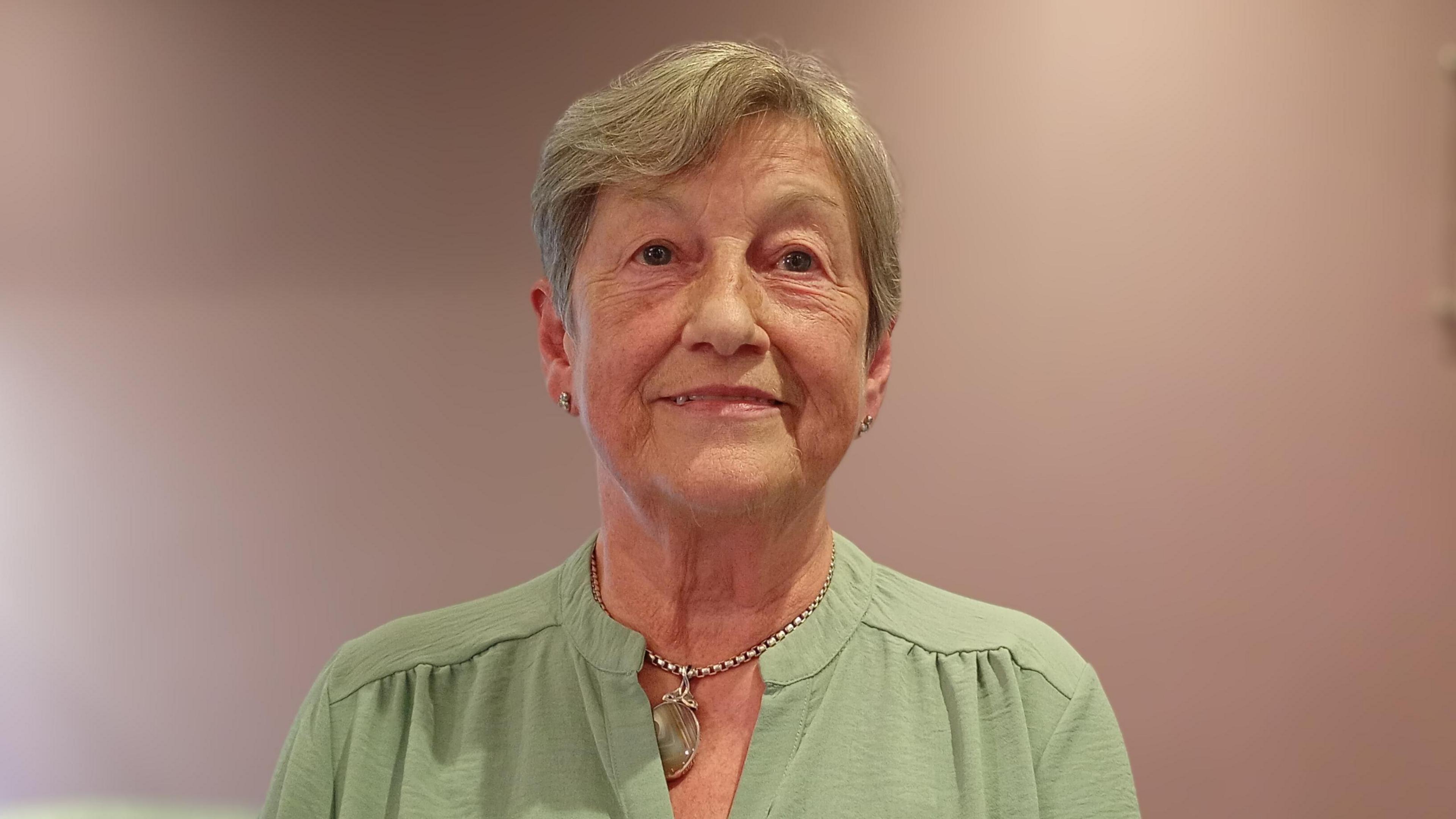 A woman, with short grey hair, wearing a green top and a necklace.