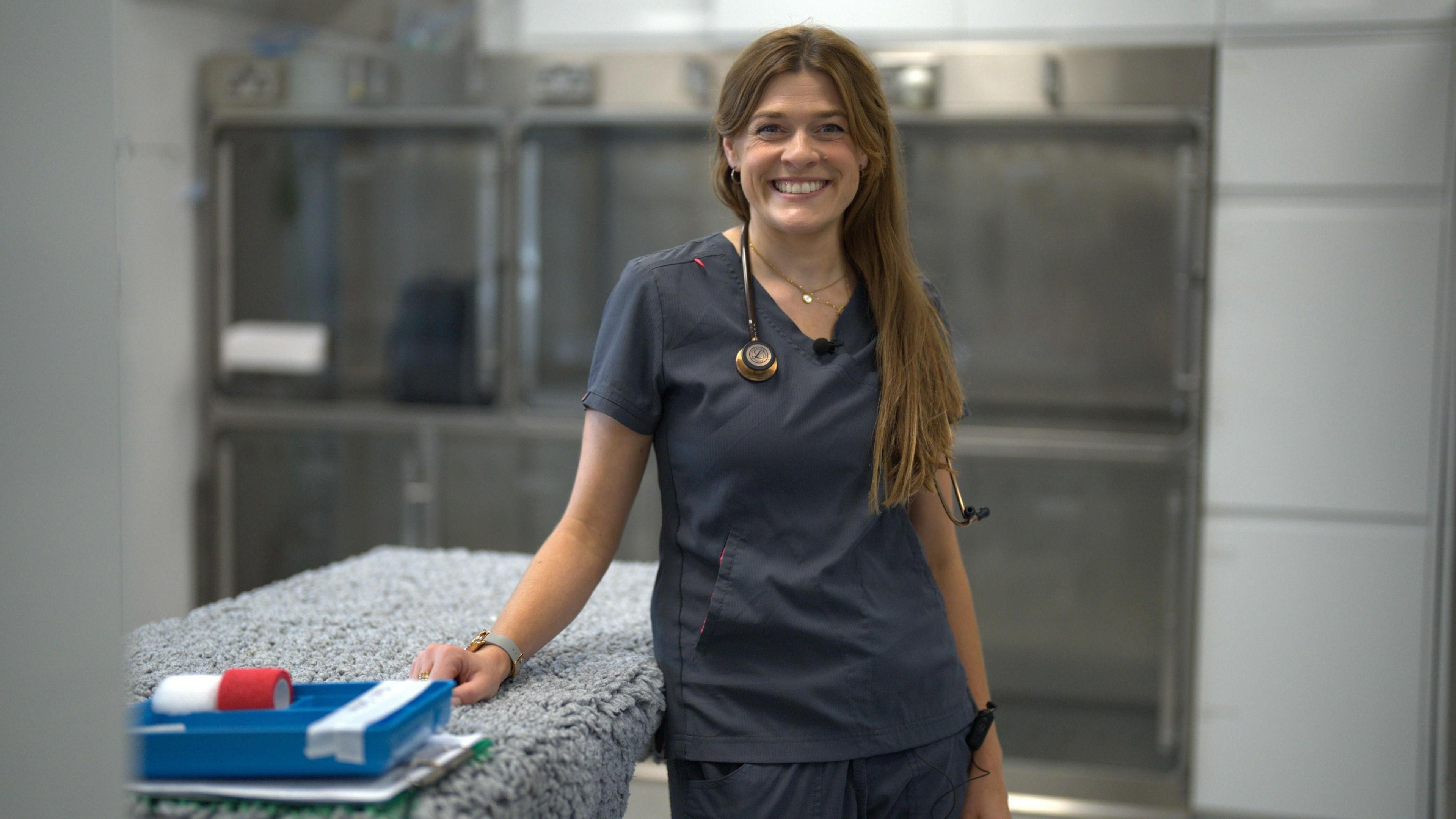 A woman with long brown hair stands next to a vet operating table that is covered in a grey fleece. She is wearing dark grey scrubs and has a stethoscope around her neck.