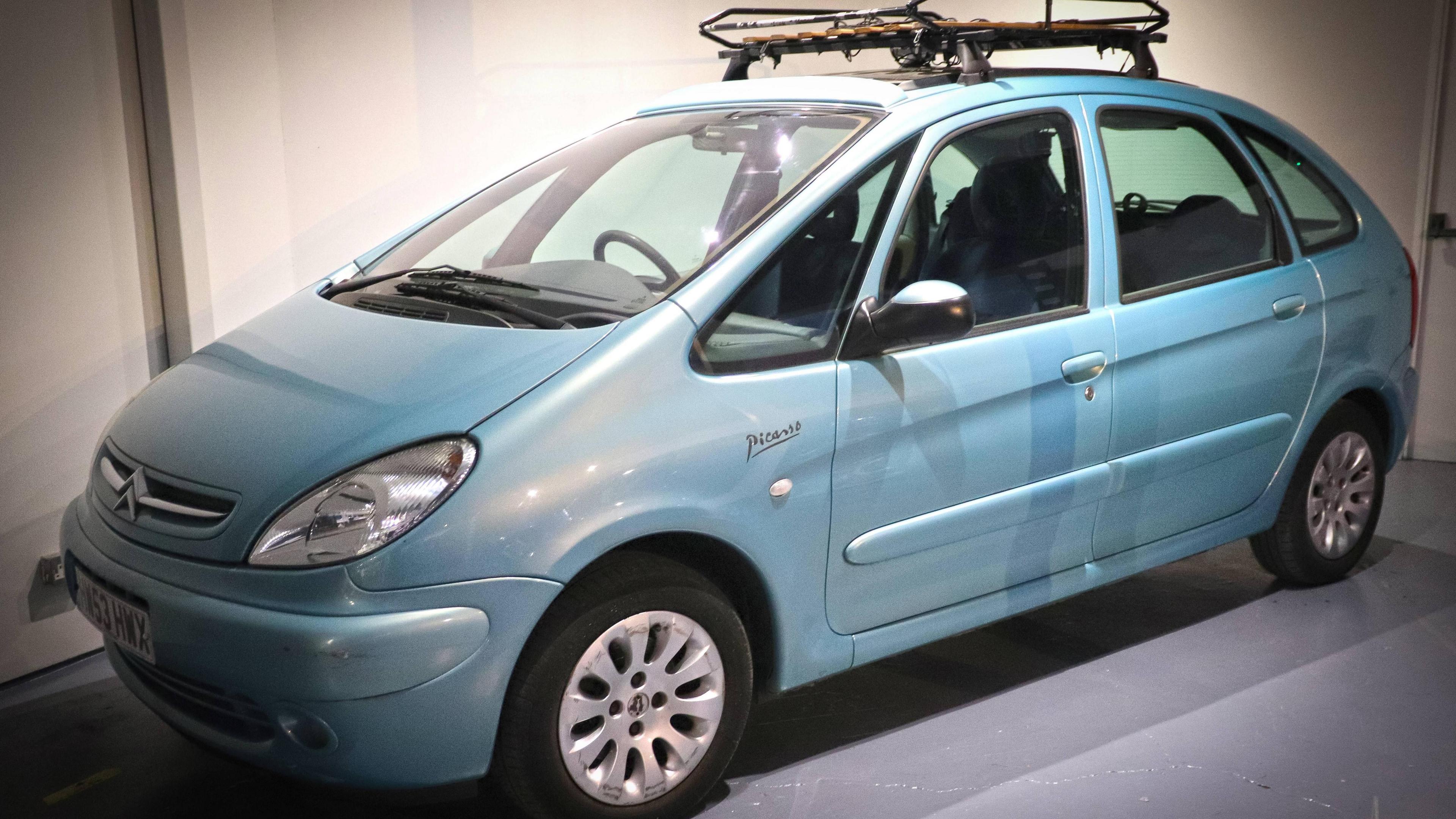 A light blue Citroen Picasso is parked next to a white wall