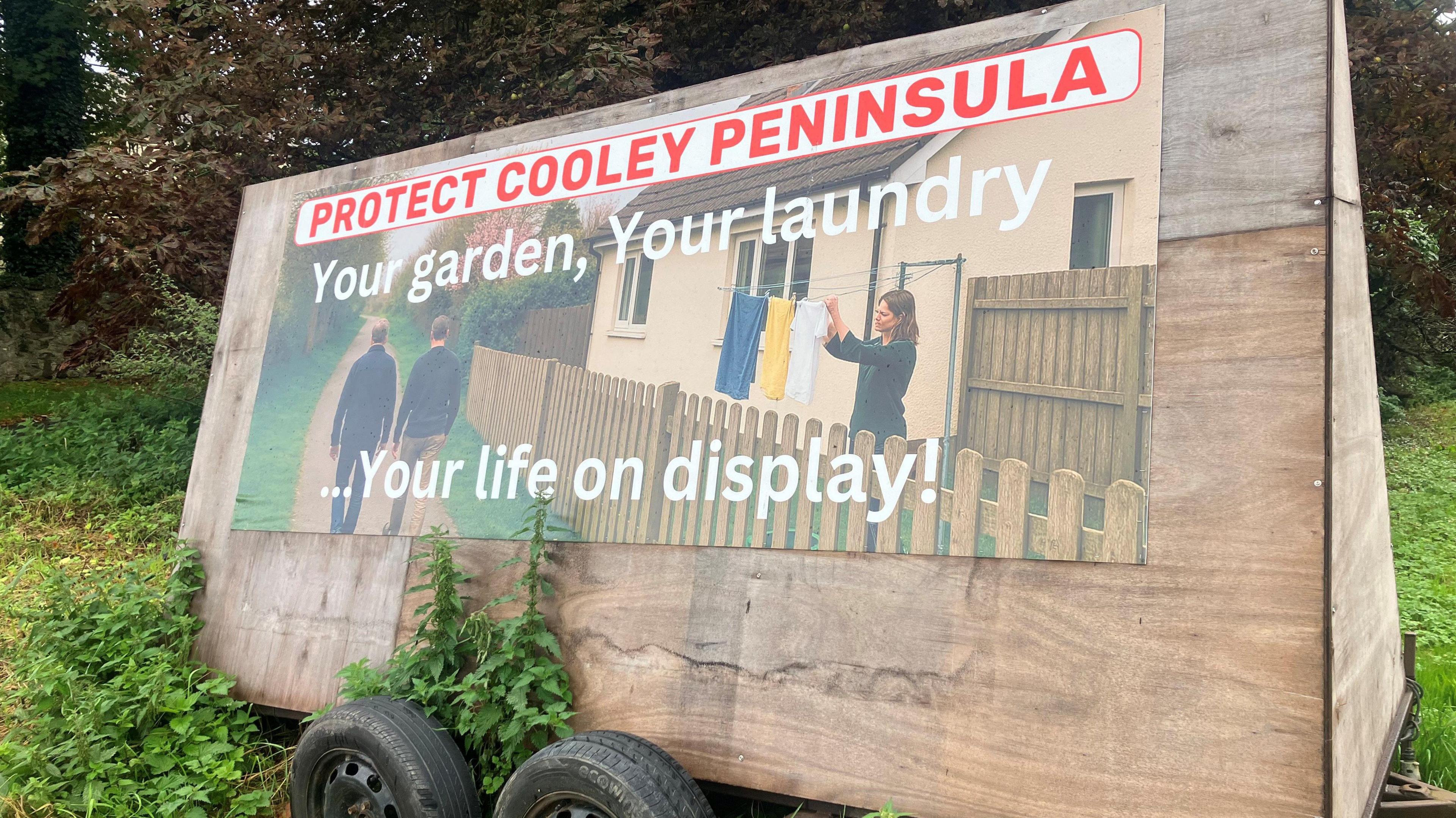 A poster says protect Cooley Peninsula. Your garden, your laundry. Your life on display. It's attached to a mobile board. It's parked on grass with trees in the background.