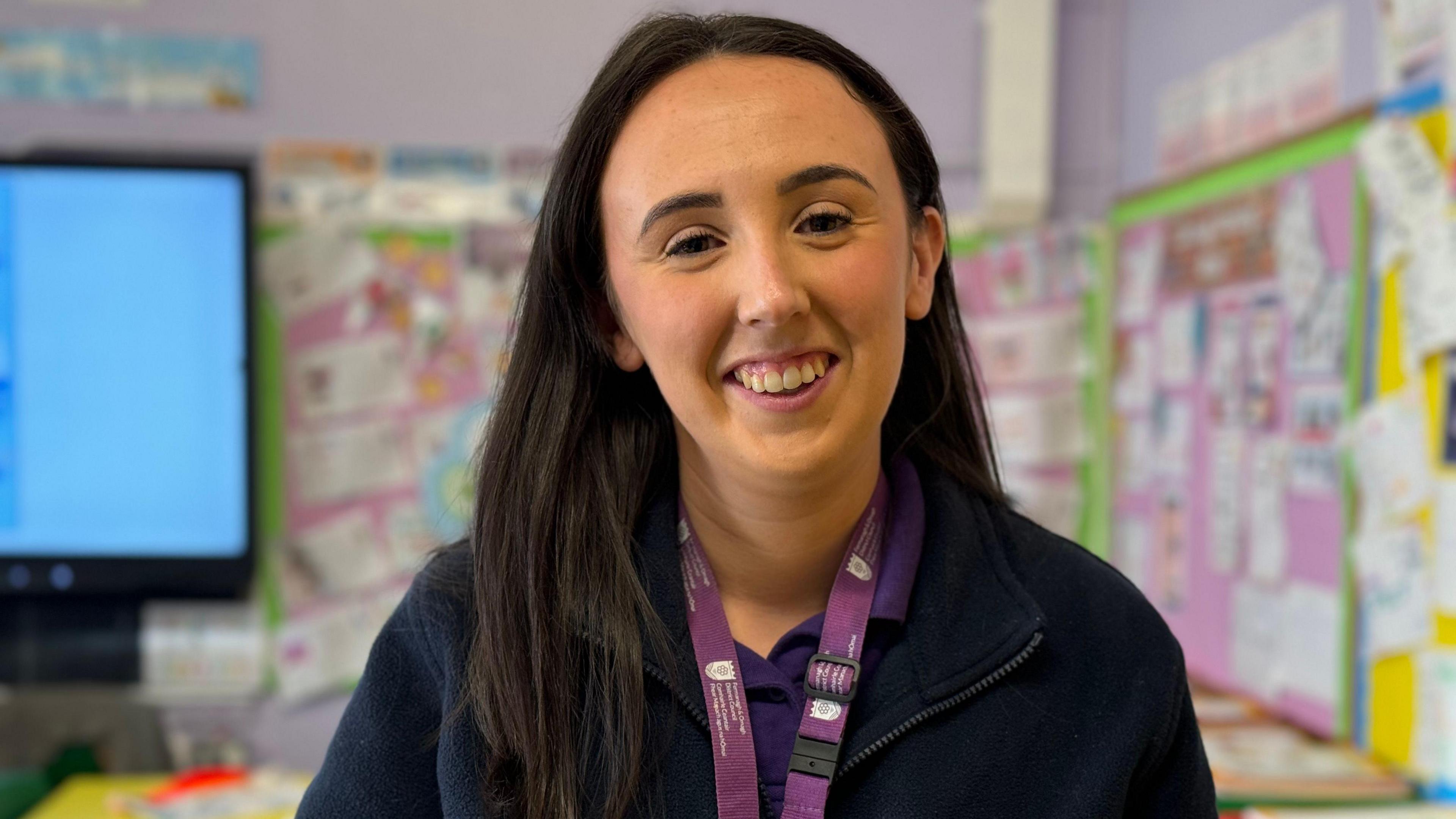 Amy Gallagher - a woman with long, dark hair wears a dark, zip up jacket and a purple lanyard. She is smiling at the camera and is standing in the middle of a classroom with wall displays and a large screen in the background.