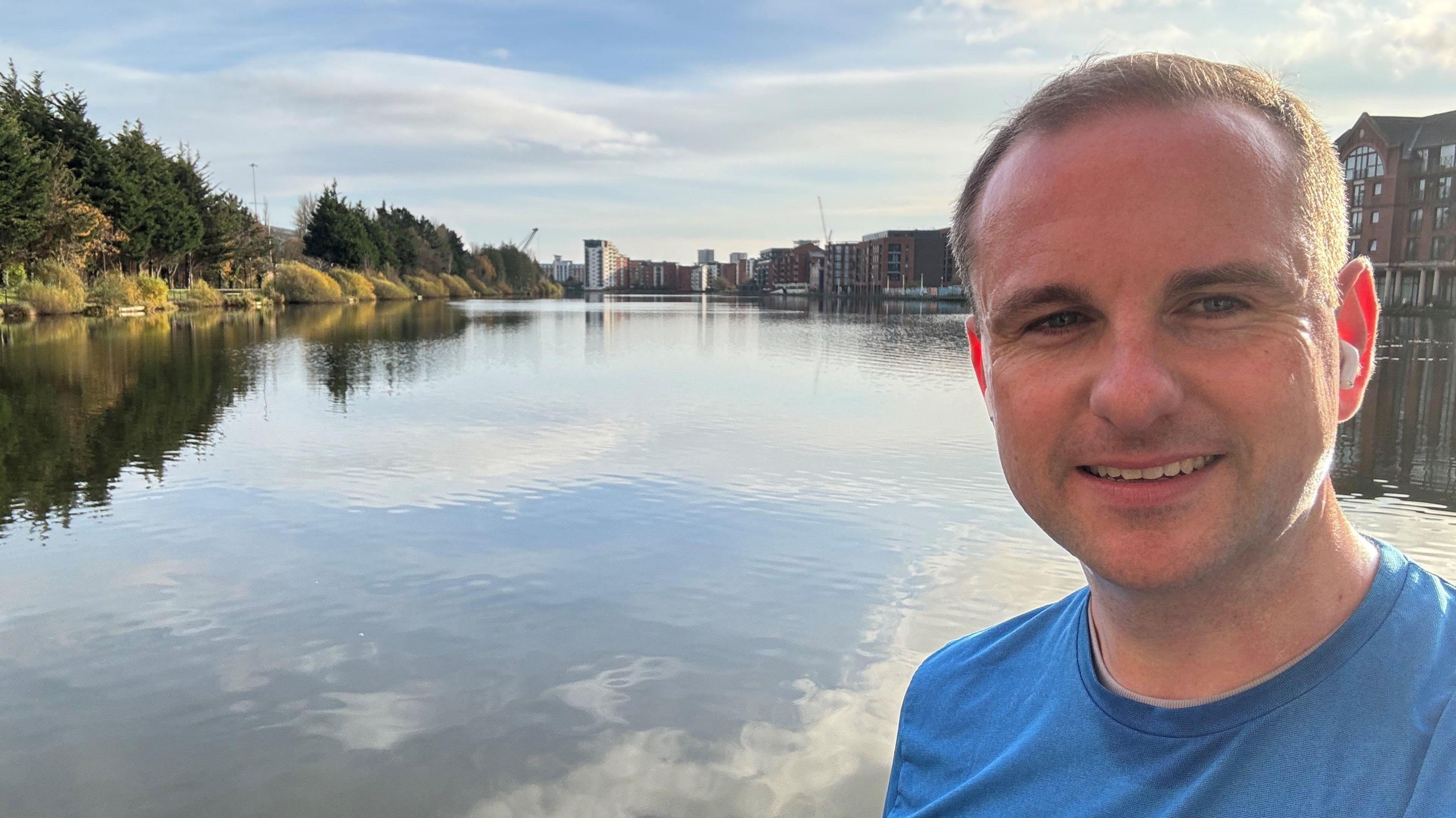 Selfie taken by Andrew Wilson in Cardiff.   He has short, fair hair and is wearing a blue t-shirt.  There is a waterway behind him, lined by tall buildings.