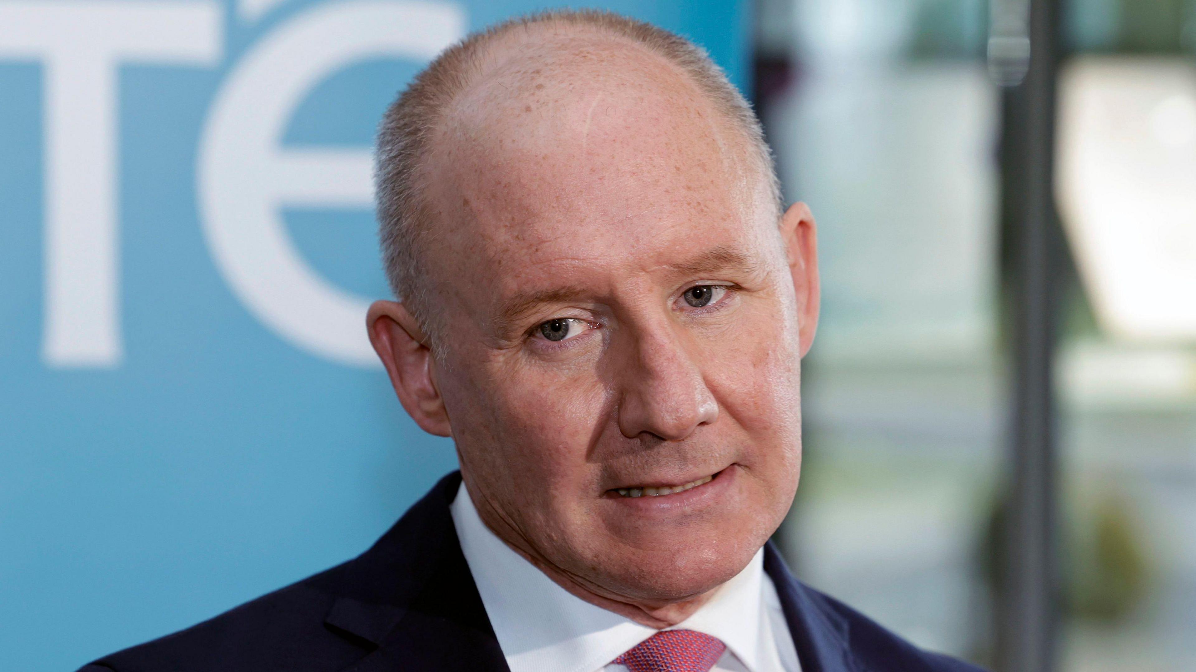 A close-up of Jim Gavin. He is an older man, with balding/short grey hair. He is mid-speech and is standing in front of a large blue and white RTÉ board. He is wearing a dark suit jacket, a white collared shirt and a pink tie.