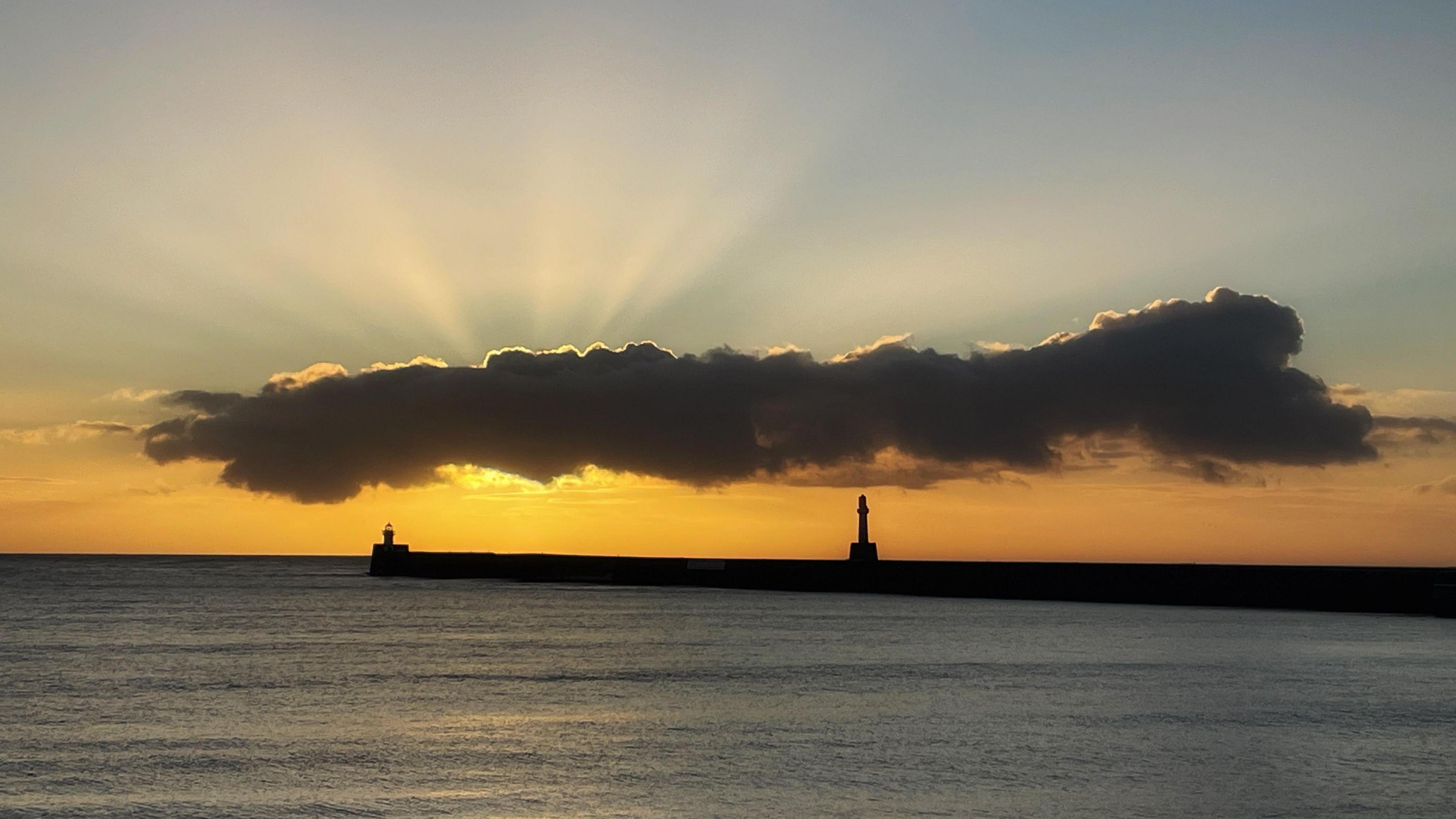 The sun is behind a long, lone cloud above a jetty jutting out to sea. Beams of light are radiating from behind the cloud.