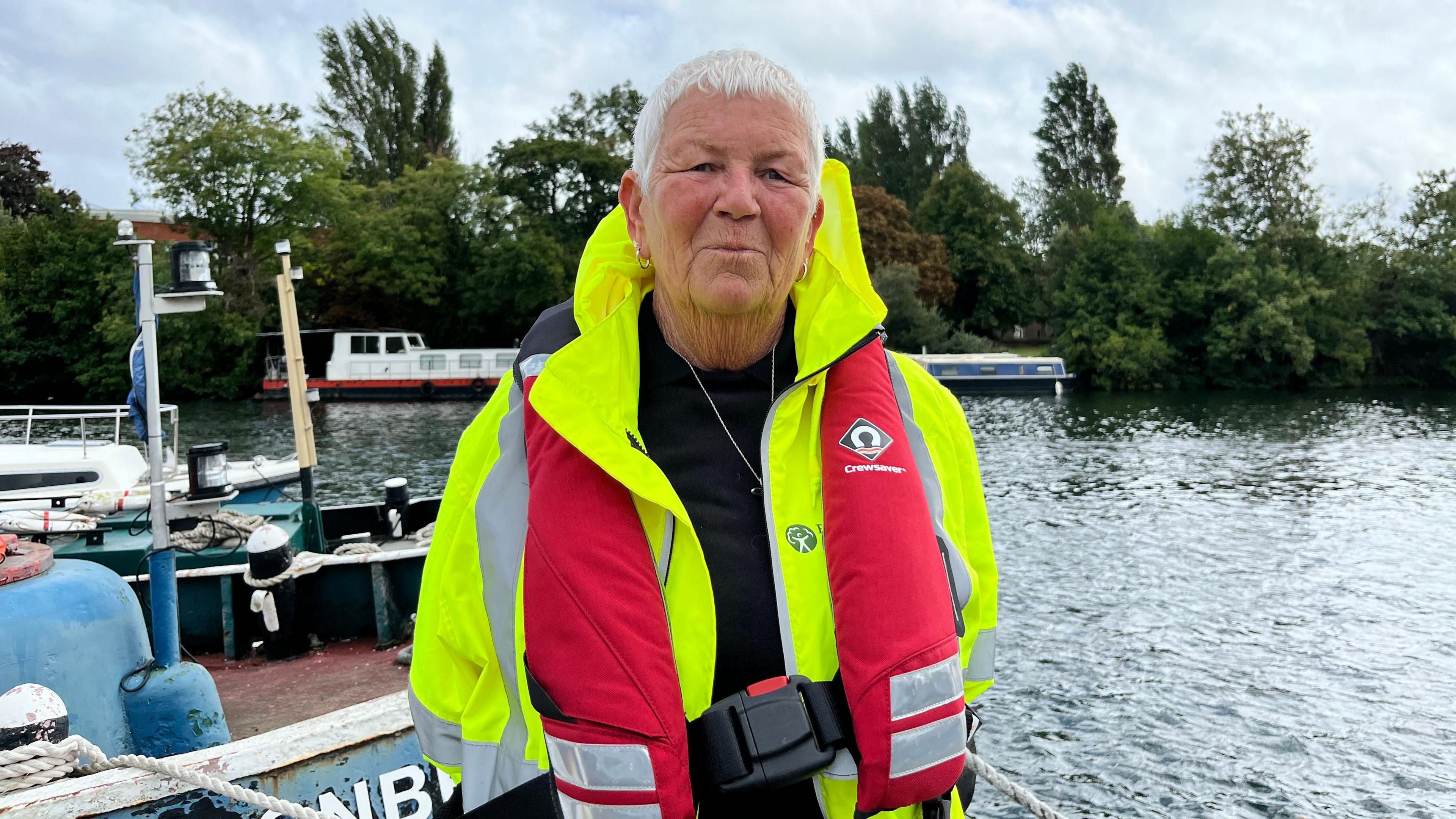 Maria Herlihy looks at the camera and smiles. She is wearing a hi-viz jacket and a red life jacket. She is standing on the edge of the river with boats and trees seen behind her and a cloudy sky.