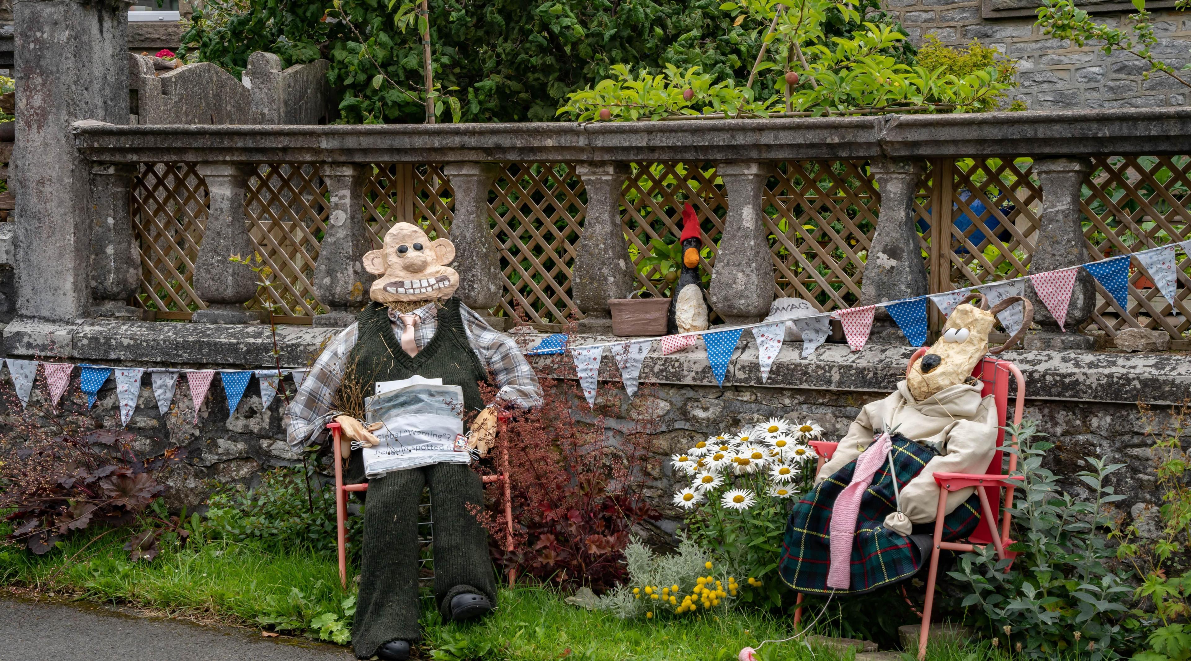 Two scarecrows depicting Wallace and Gromit sit in deck chairs on a grass verge in front of a stone wall. 