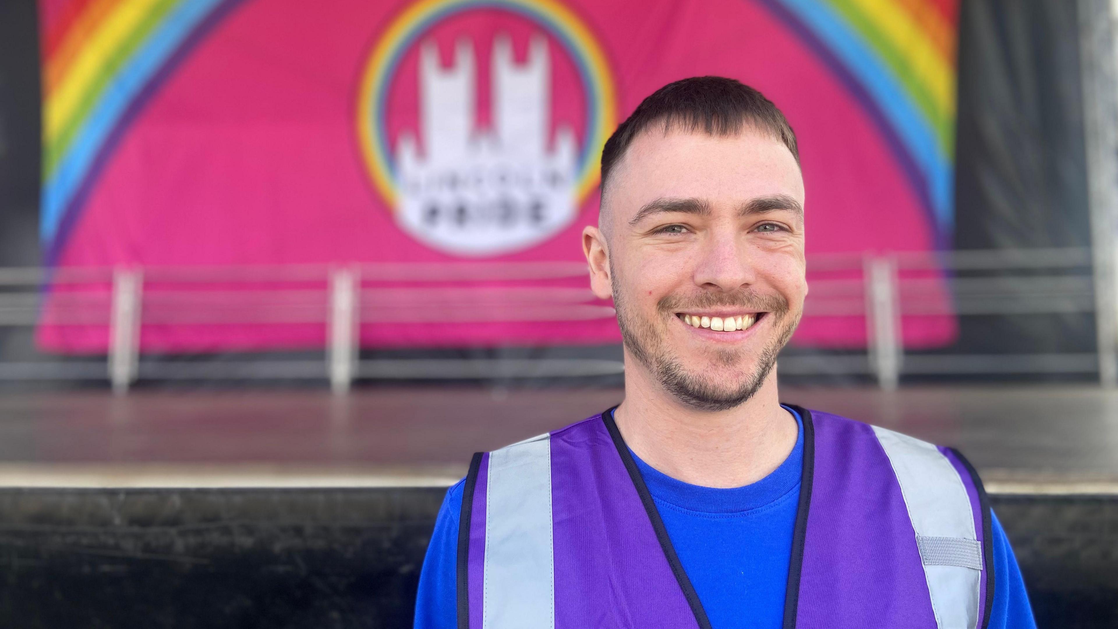 Jason Reid has short dark hair and a close-cropped beard, he is smiling in a purple high-vis vest in front of the main stage at Lincoln Pride.