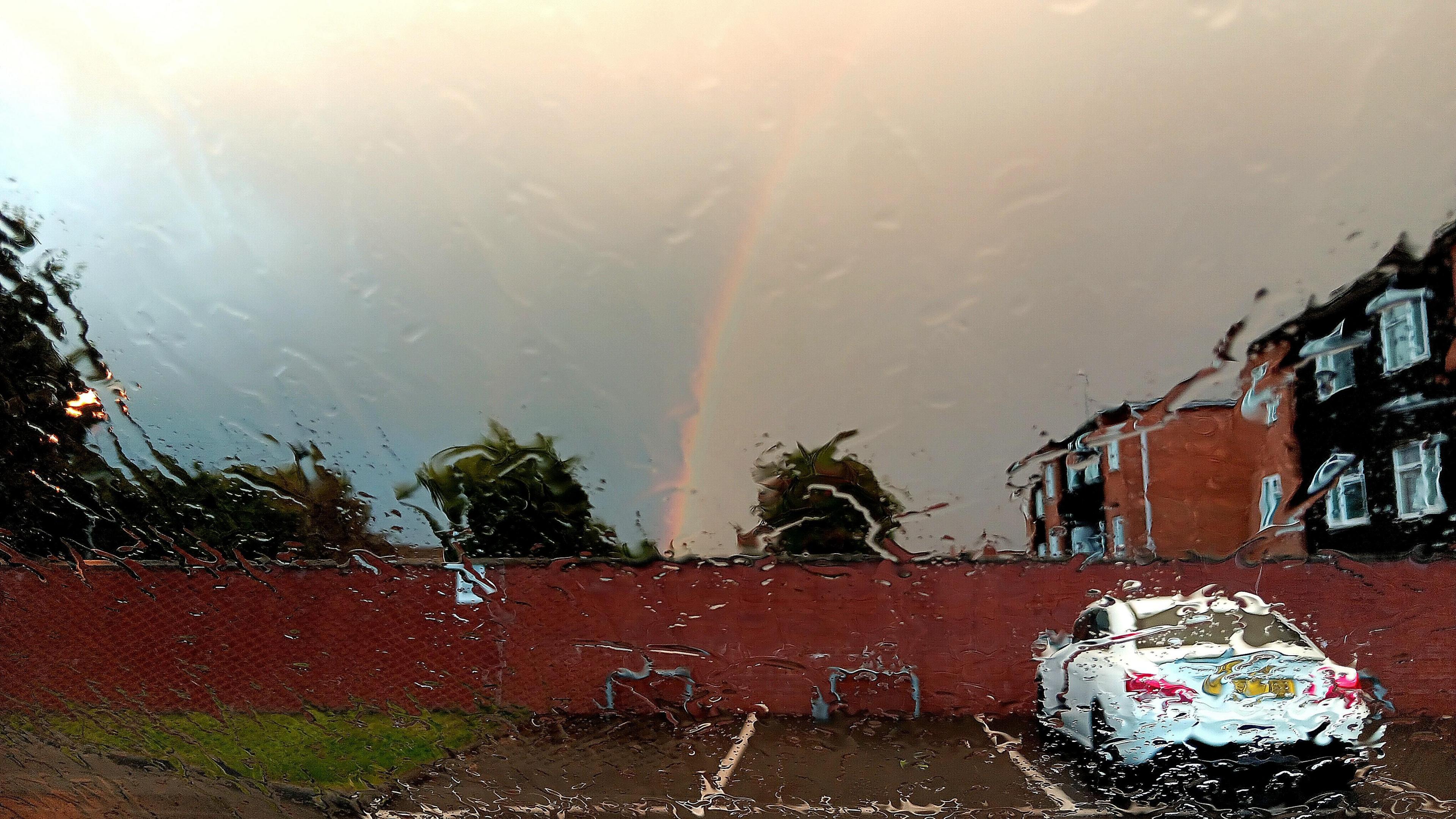 The picture is taken through wet glass, distorted by raindrops. It shows a parking area outside a house with a white car parked and more houses to the right. Straight ahead is a red-brick wall with a rainbow dissecting the dark grey sky above.