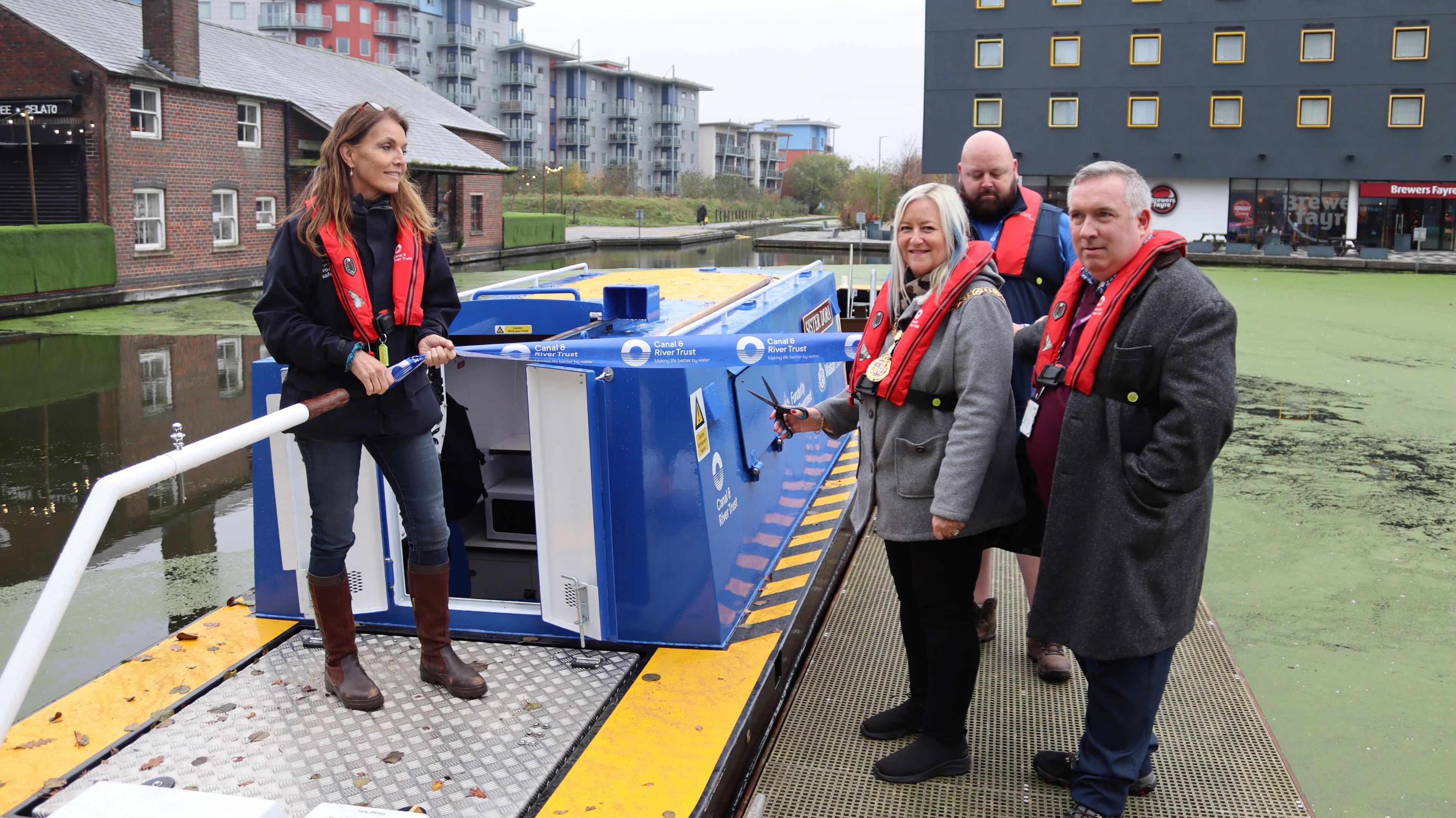 Two men and a woman stand next to a canal boat wearing red life jackets. The woman is holding a pair of scissors. On the canal is a woman holding the boat's tiller.  A blue ribbon is stretched out between the woman and one of the men as the other woman is about to cut it. The boat is sitting on the water, mostly covered in green, with buildings visible in the background.