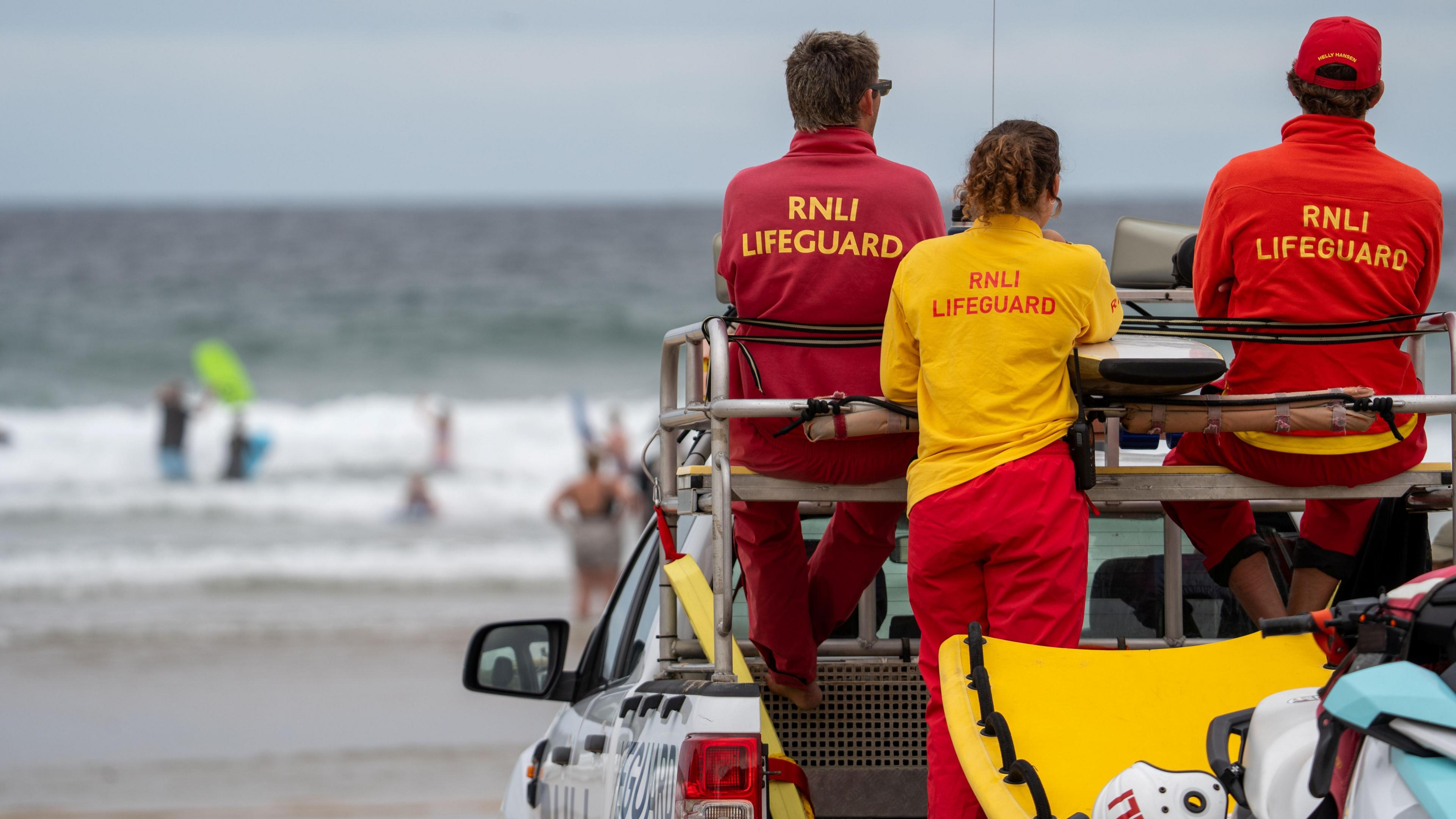 Two male lifeguards are sitting on the roof of a pick-up truck and and one female lifeguard is standing in its flatbed. They are in branded uniform and are looking towards the sea. The sea is blurry as are the swimmers and body boarders in its. 