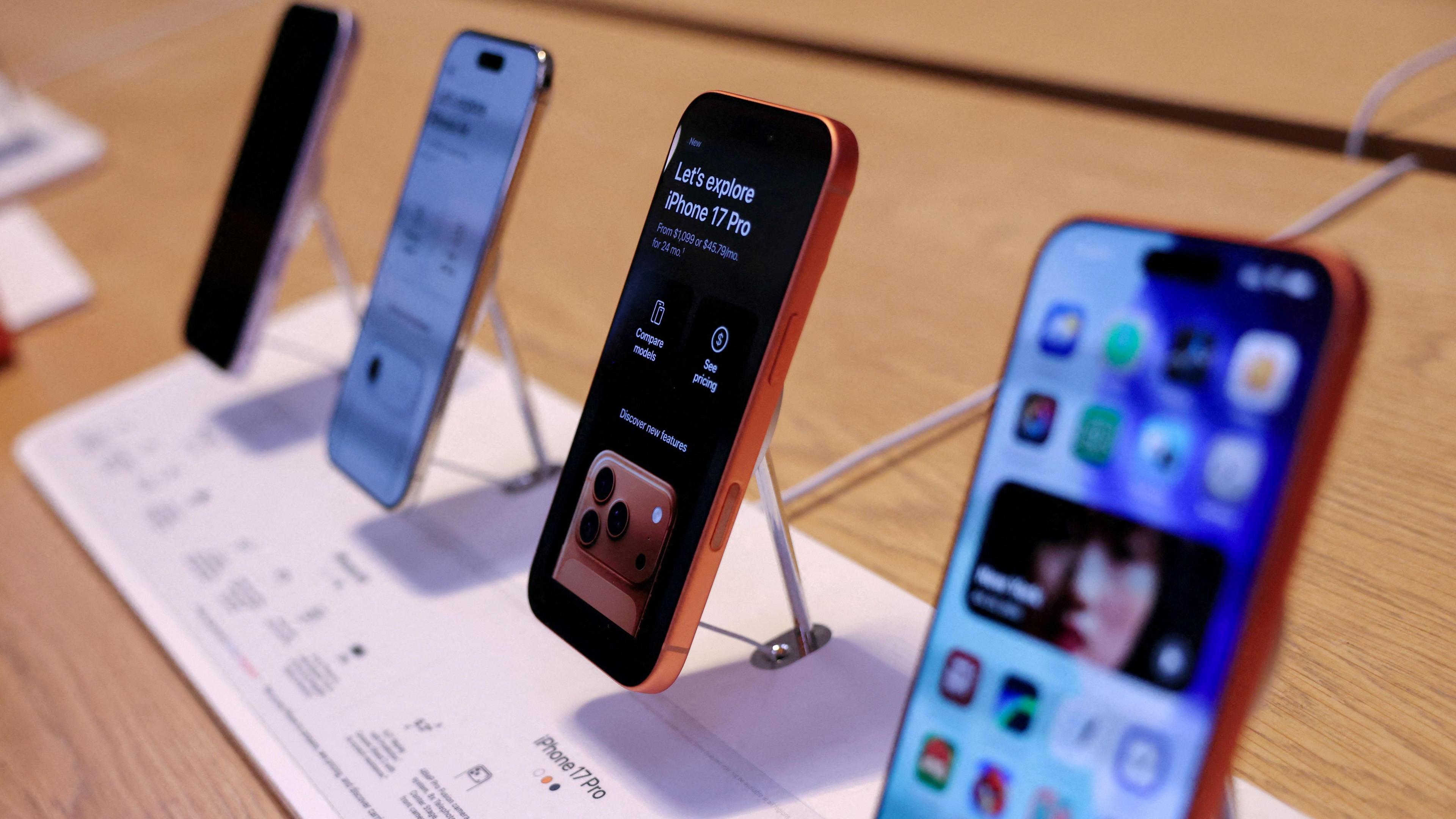 Four iPhone 17s on display in an Apple store. The phones are lined up in a display on a wooden shelf. 