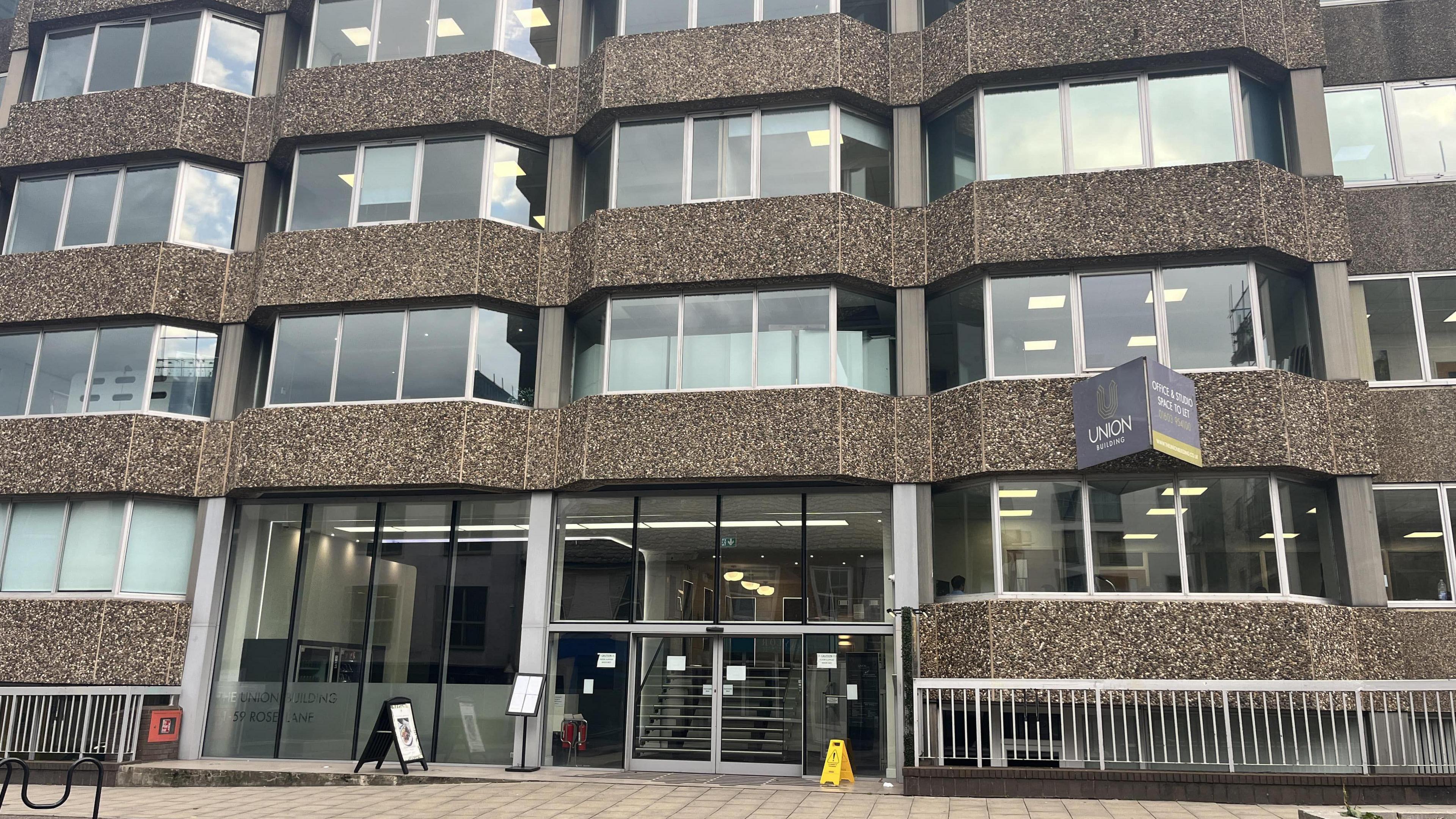 Front aspect of a large office building showing entrance and windows. The pavement can be seen alongside signage. 