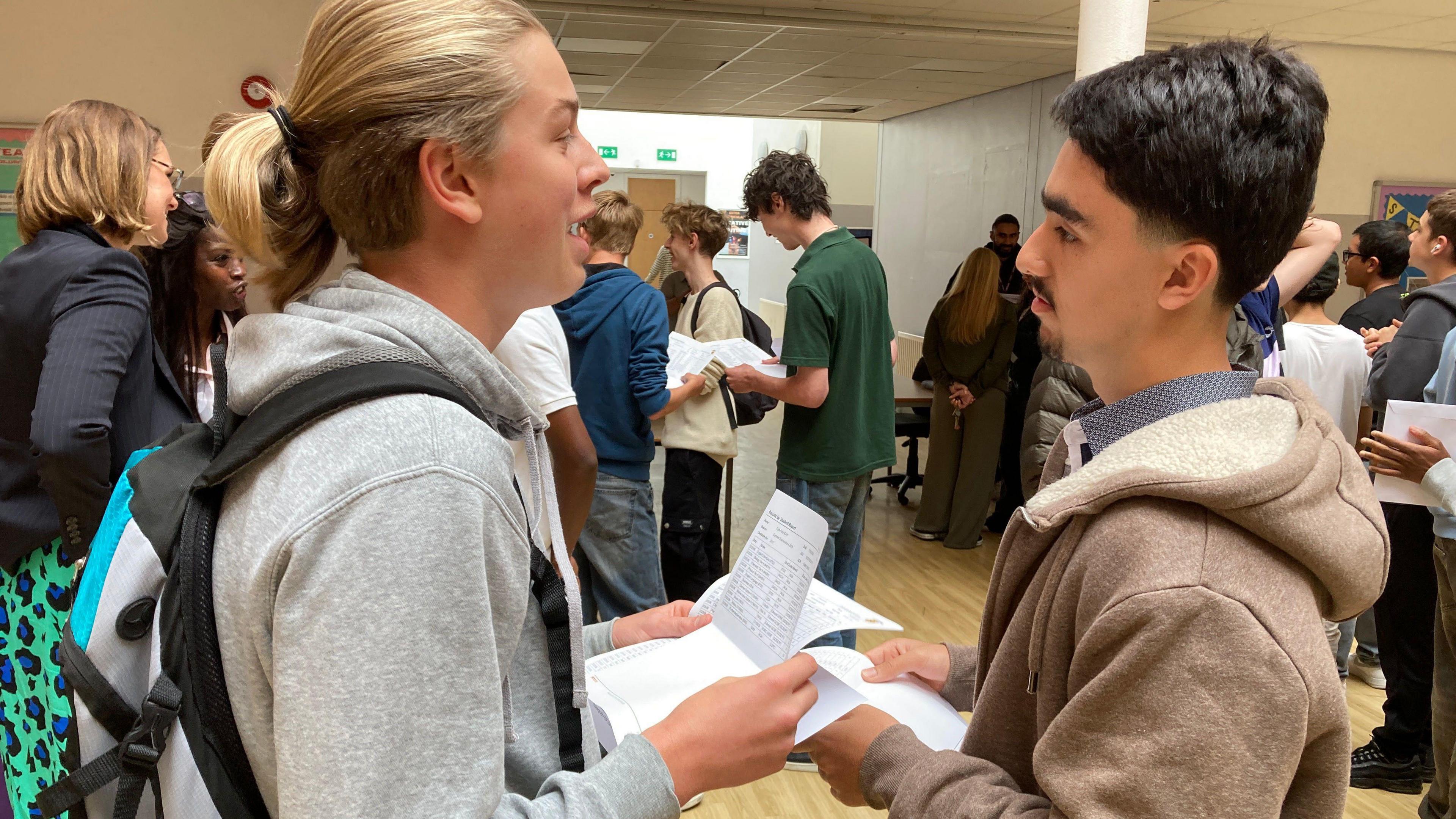 Bilal stands to the right, talking to his friend opposite him on the left of the image. Bilal has short black hair and a short beard. He wears a brown hoodie, with a blue print shirt underneath. His friend has blonde hair tied back into a ponytail and a light grey hoodie. There are students all talking and gathered in the background in their school hall.