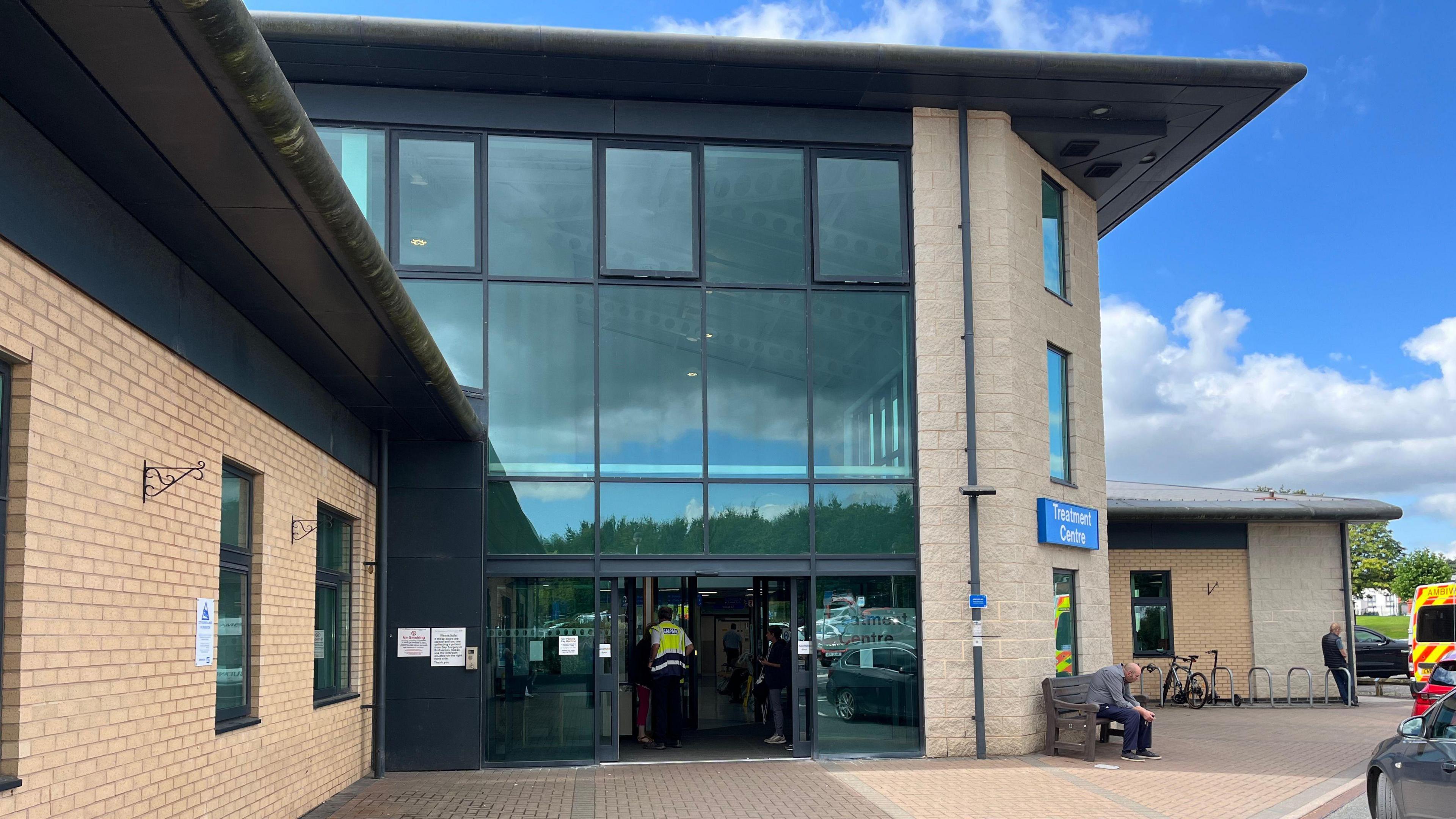 The side of a hospital entrance building. It is partially light brown brick with floor to ceiling glass windows on the side. A worker is walking in the door and a man is sat on a bench outside.