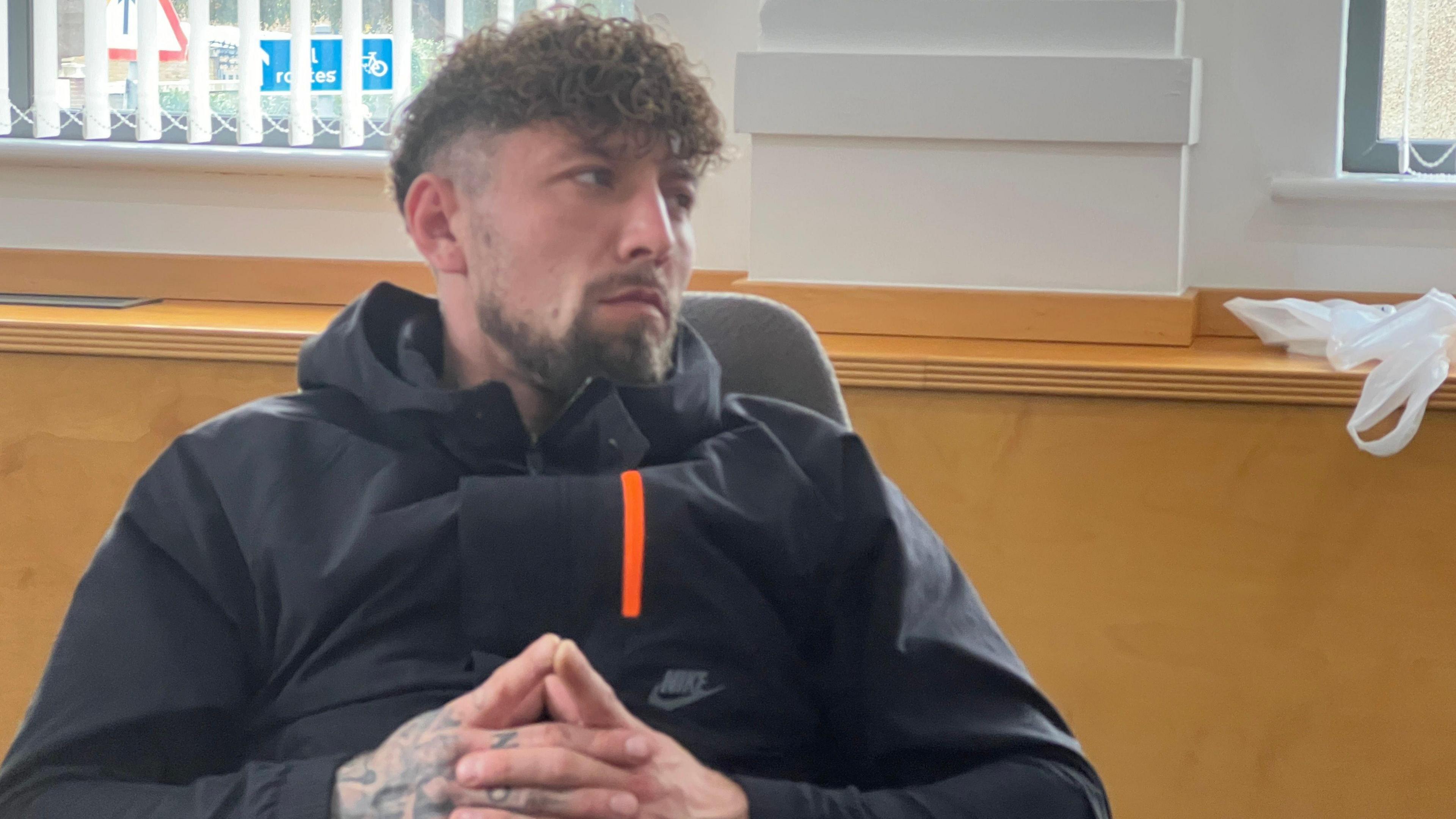 Dean Plunkett is slouched in his chair, wearing a Nike jacket. He has his head turned towards the right, watching the screen. His hands tattooed and clasped together and resting on his stomach. 