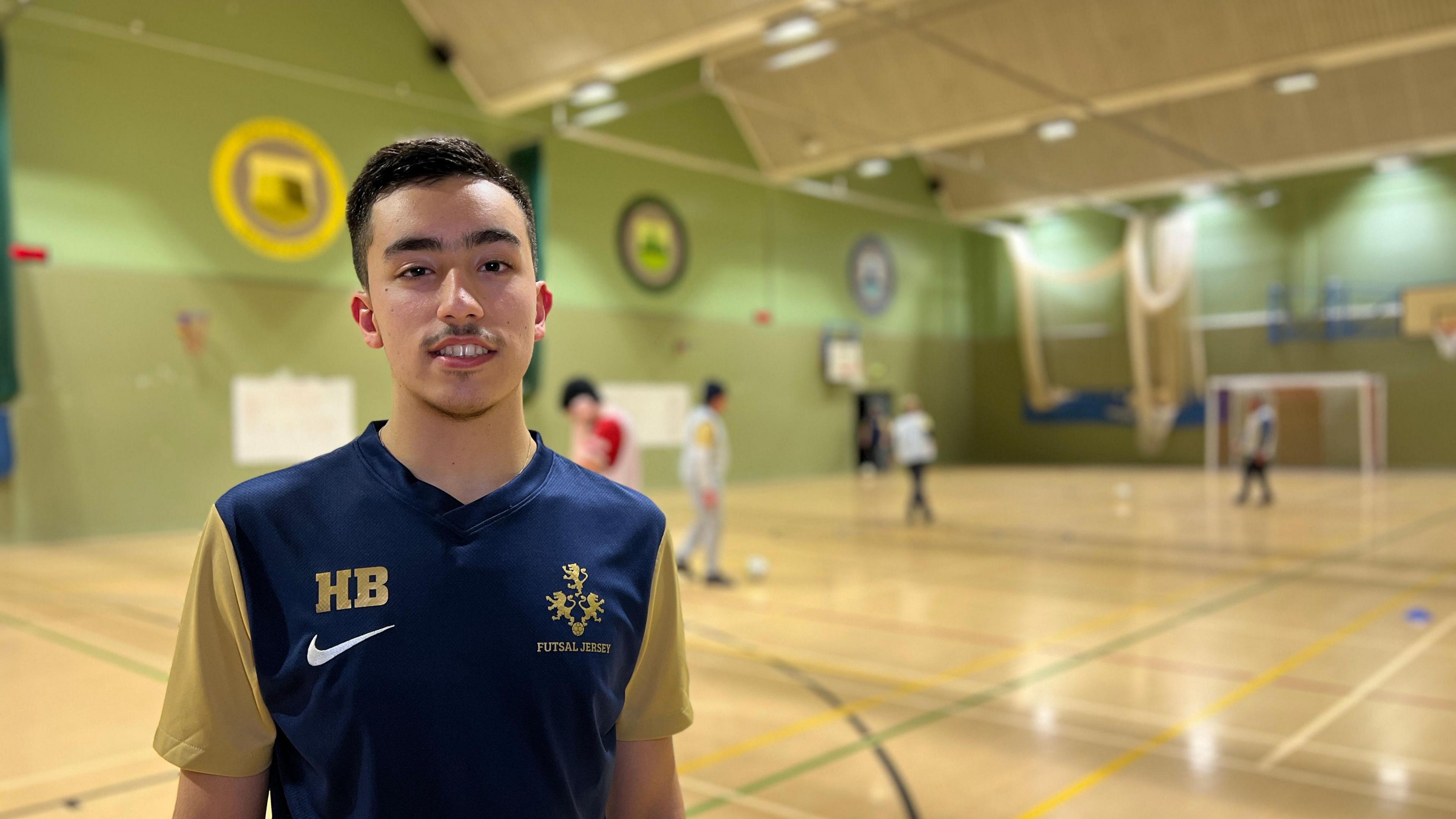 Humair looks at the camera while people play futsal behind him. He's wearing a blue football top with yellow sleeves and he has short black hair which has been shaved at the sides and a small goatee. 