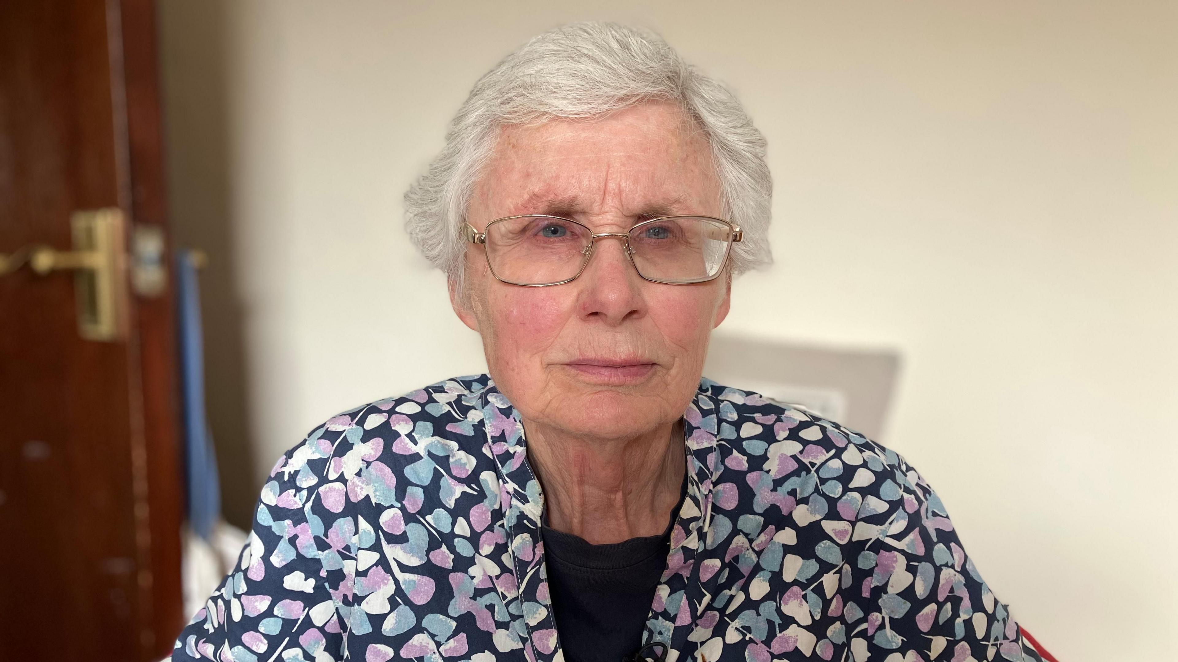 a woman in a patterned blouse sitting in front of a cream wall with a sad expression
