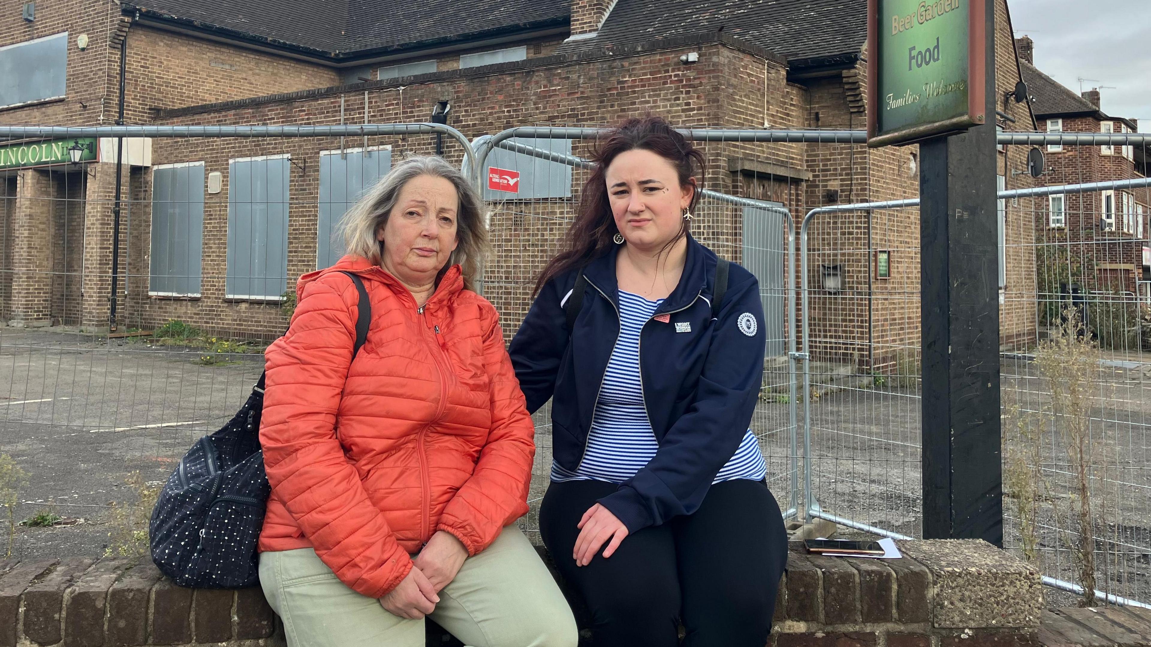 Jane Jackson (left) and EM-B (right), sitting on a wall in front of a derelict pub. Jane has light hair and wears an orange jacket and light trousers. EM-B has dark hair and wears a striped top, dark coat and dark trousers.