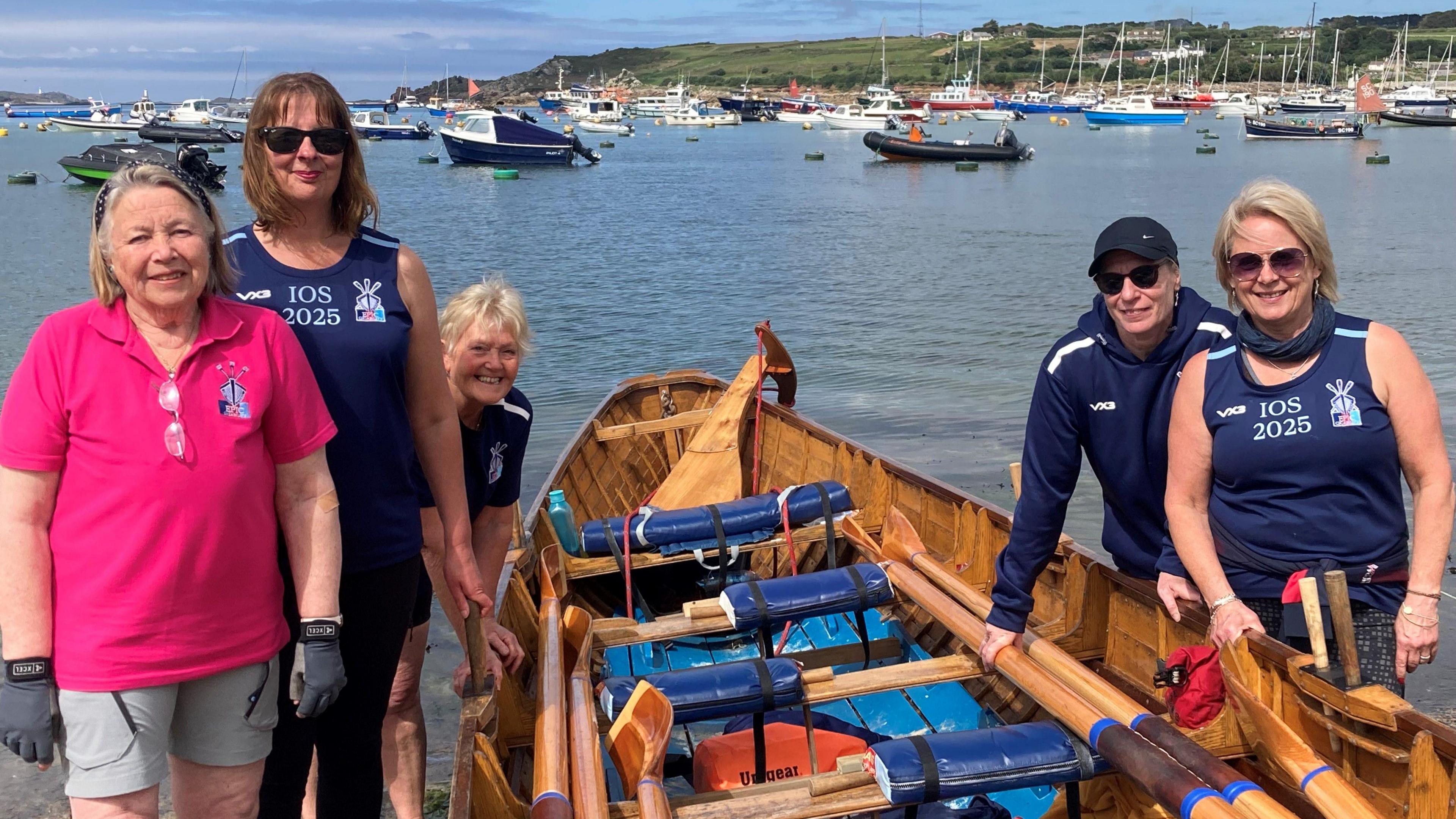 A team of rowers look at the camera while posing with a wooden gig boat in a bay
