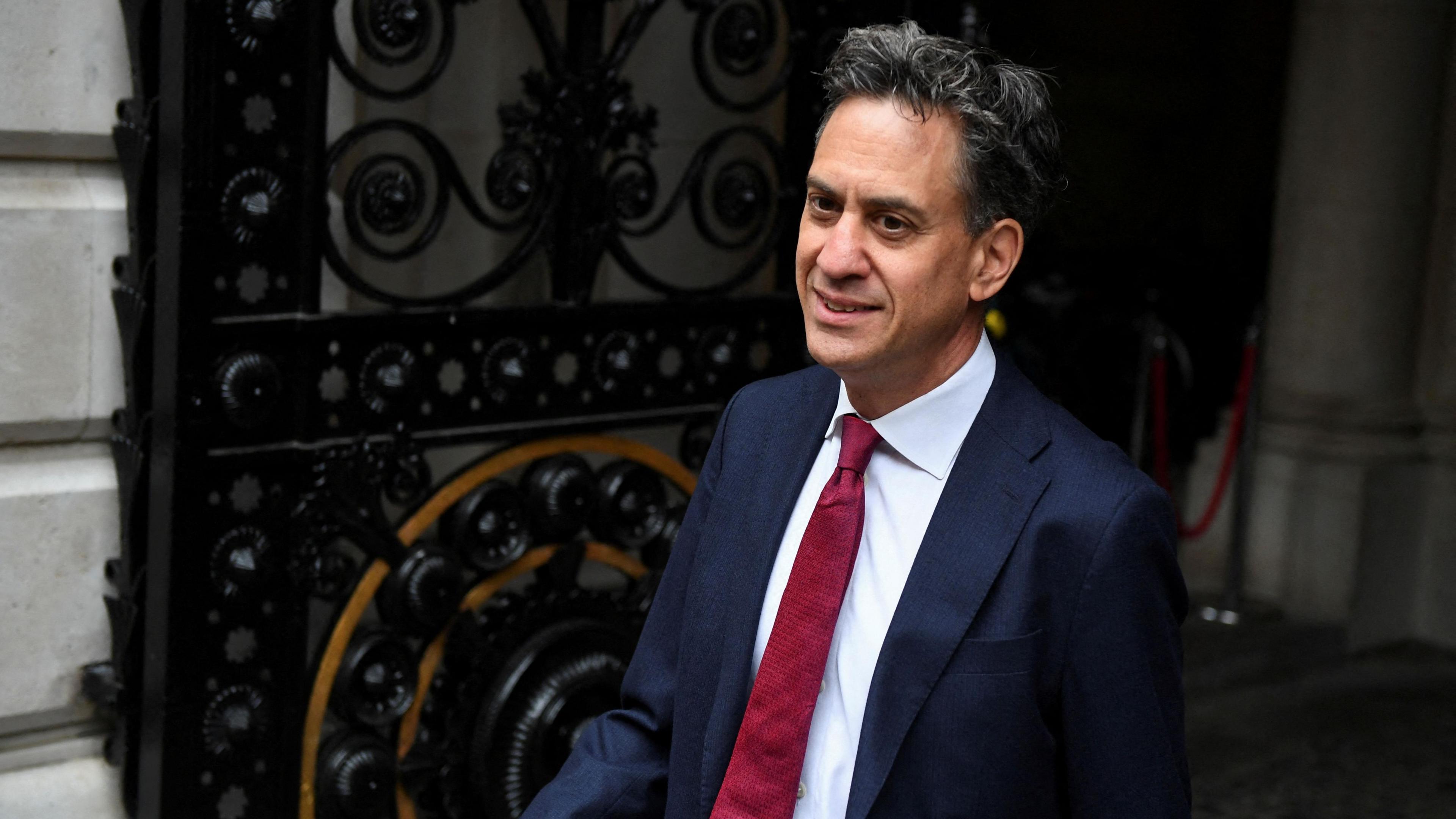 Ed Miliband walking out of a large ornately-decorated iron gate outside Downing Street in London. He is wearing a dark blue suit, white shirt and red tie.