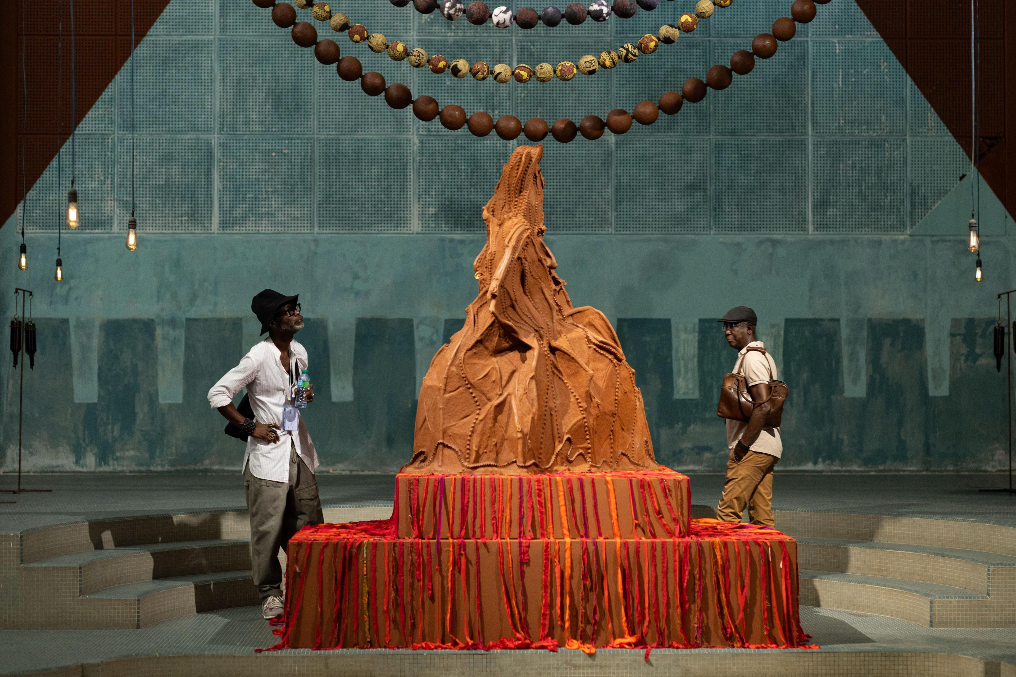 Two men look at a large rust-coloured art installation which is shaped in a peaked form.