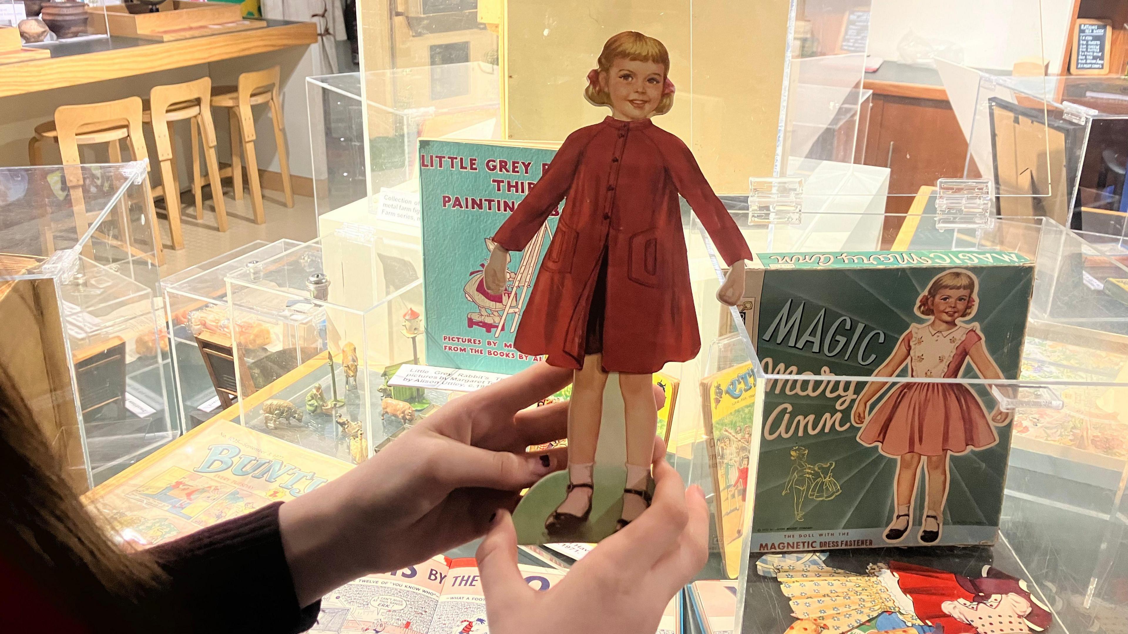 A cardboard cut-out doll is held in the hands of a girl. The doll has blonde pigtails and is wearing a red coat, black skirt and black shoes with white socks.