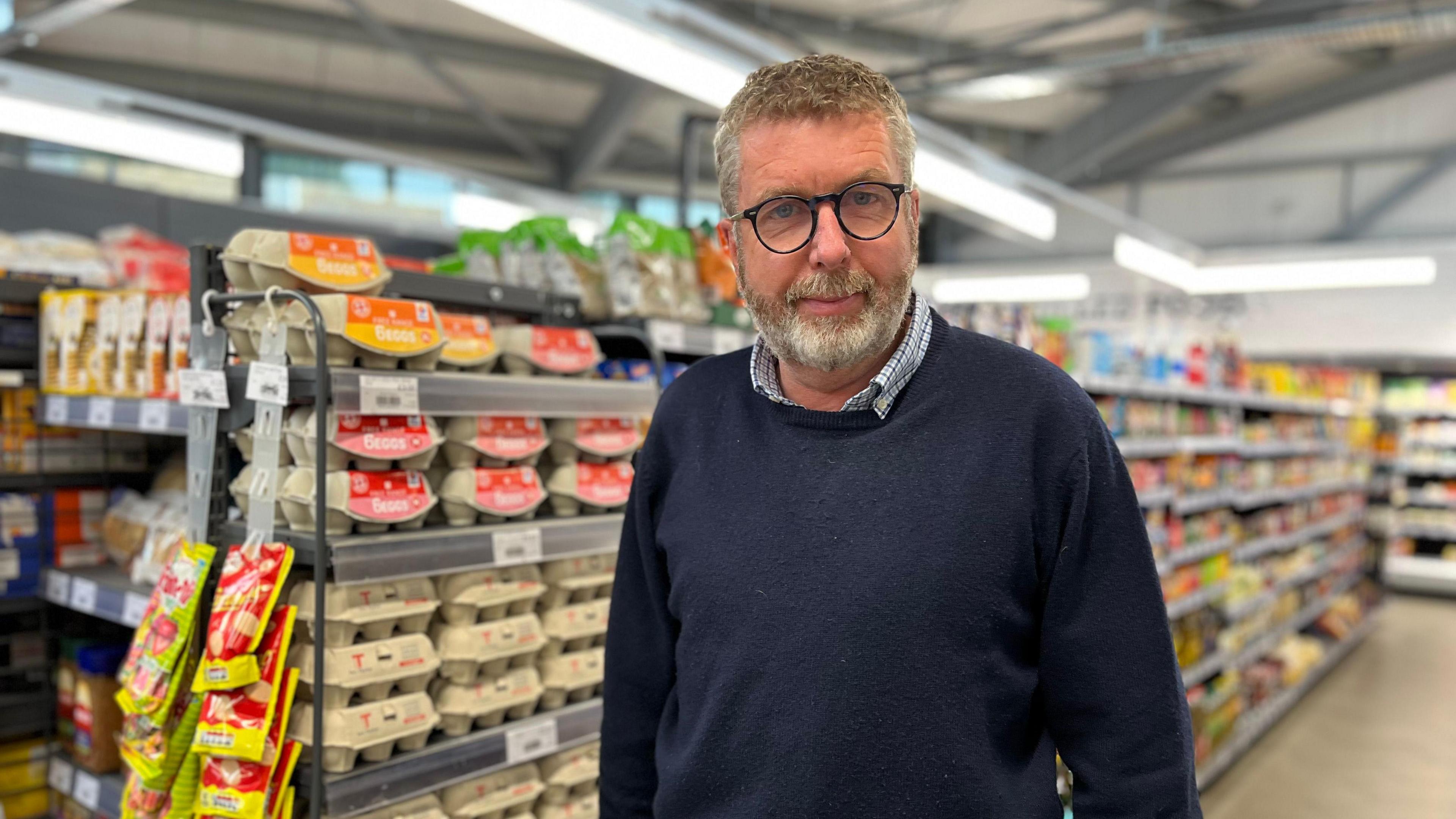 A man wearing glasses, a checked shirt and blue jumper stands in a petrol station shop in front of rows of eggs for sale.