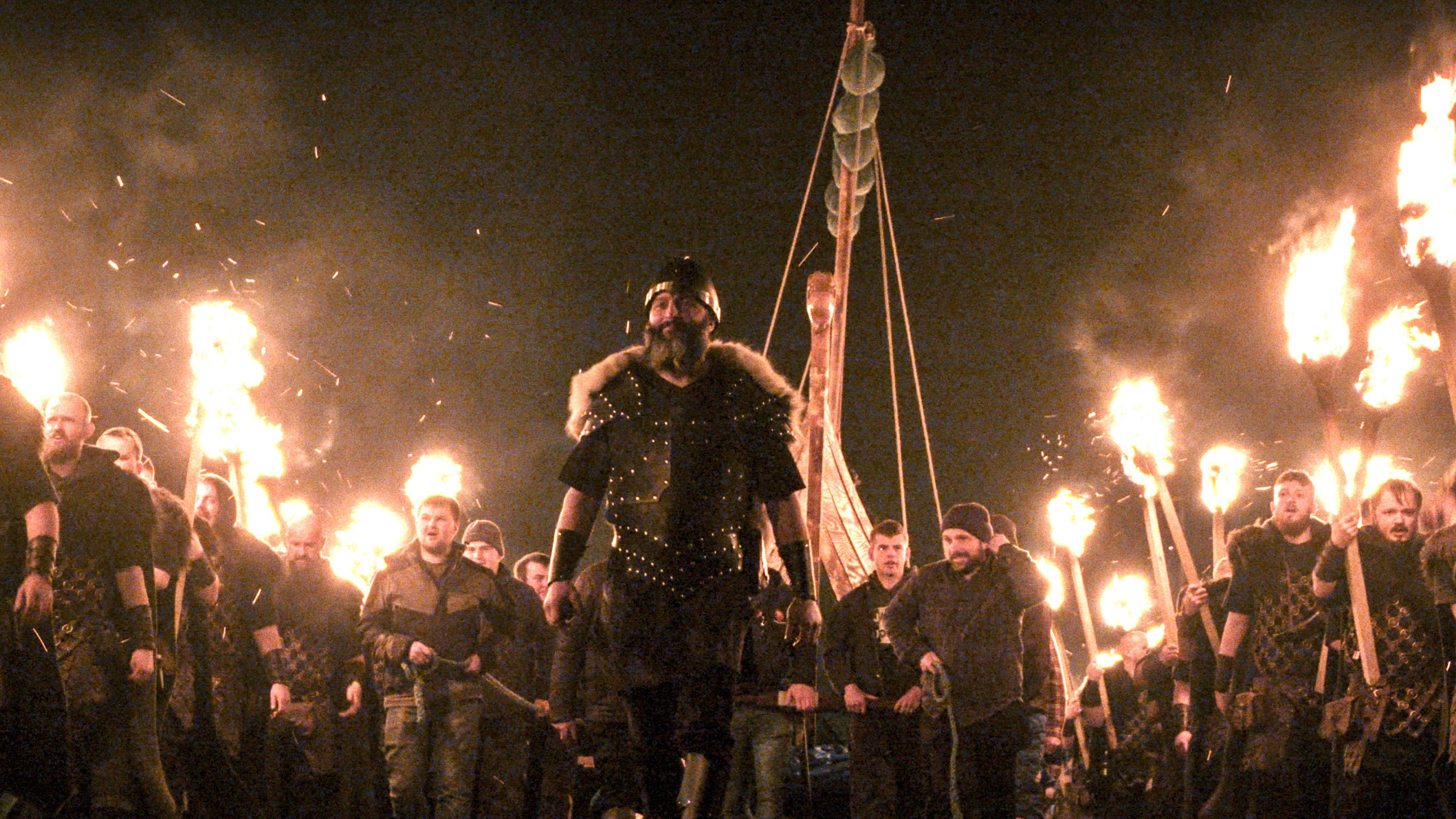 A bunch of men in Viking outfits and burning torches march in a line. Some are in modern dress too.